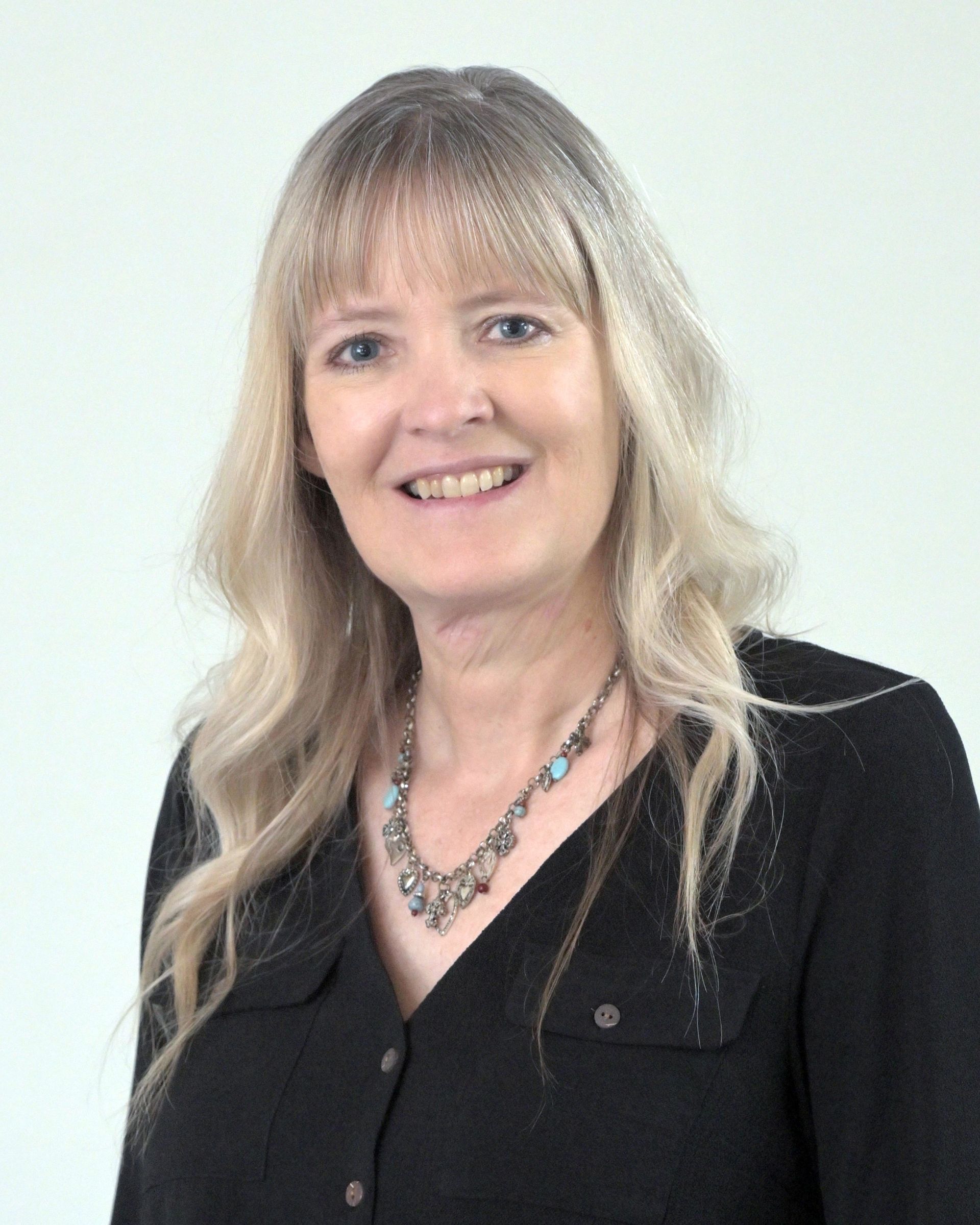 Blonde woman smiles, wearing a black turtleneck, against a blurred background.