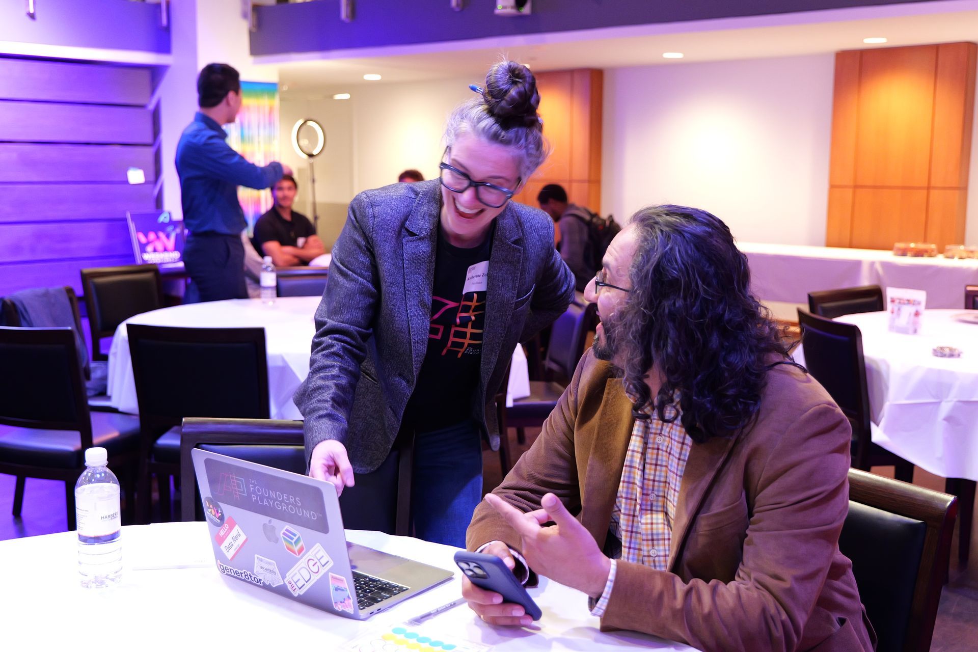 Two people at a table, one pointing at a laptop screen, the other looking at a phone. Indoor setting with tables and people.