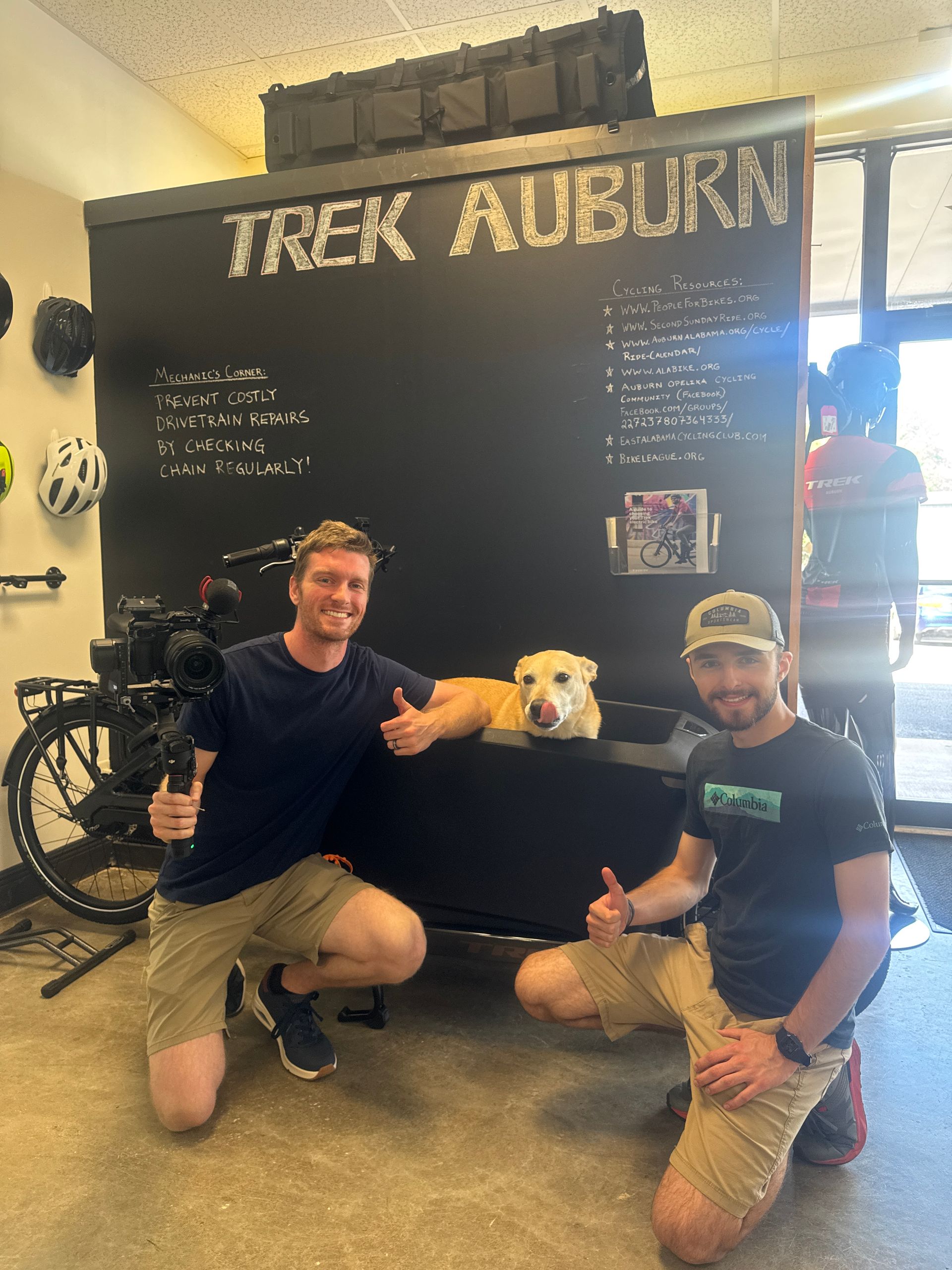 Two men and a dog pose in front of a Trek Auburn sign. The men give thumbs up, with the dog resting in the bike trailer.