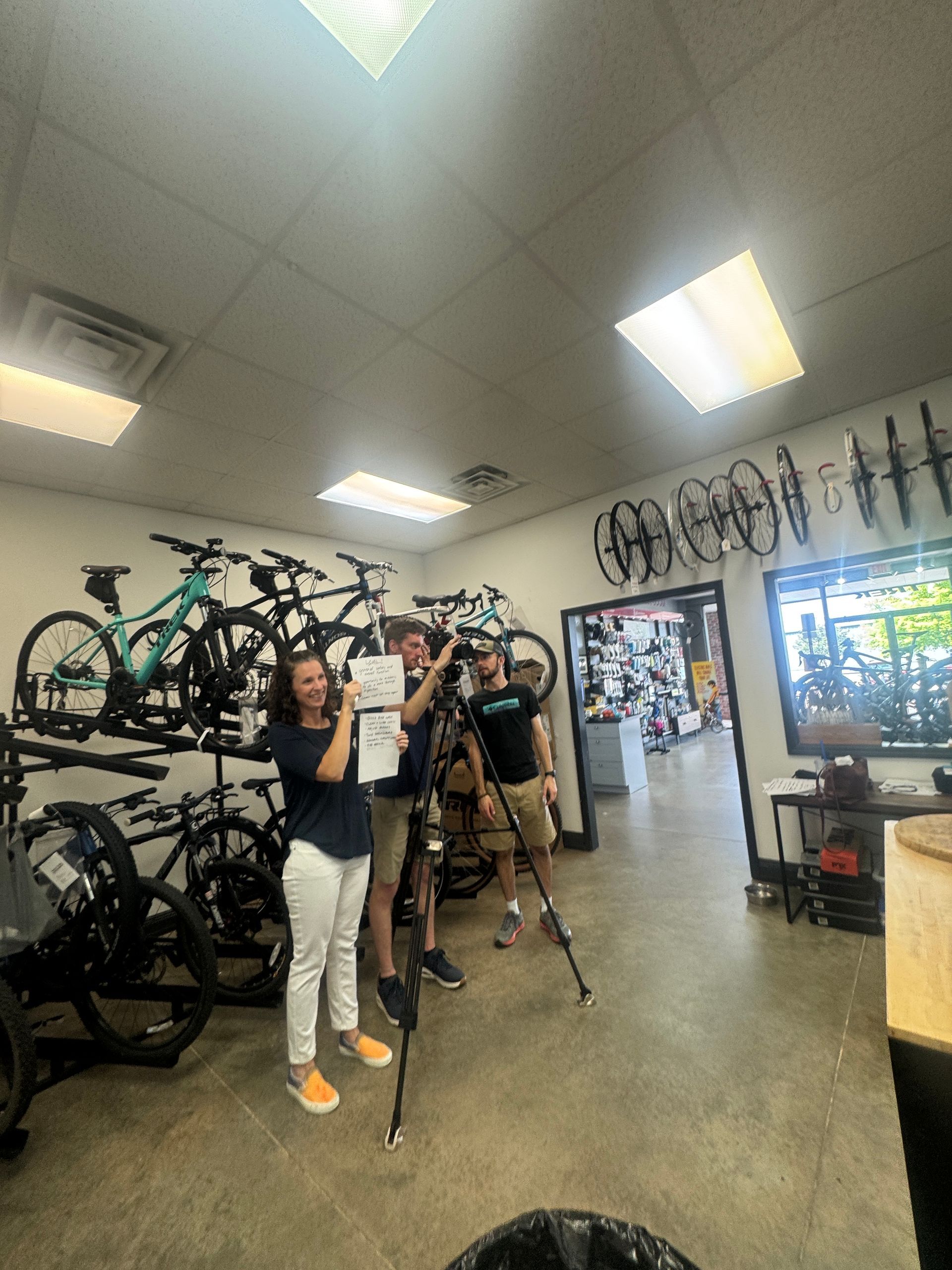 Three people in a bike shop smiling, holding a paper, and filming. Bikes hang on the wall and in the background.