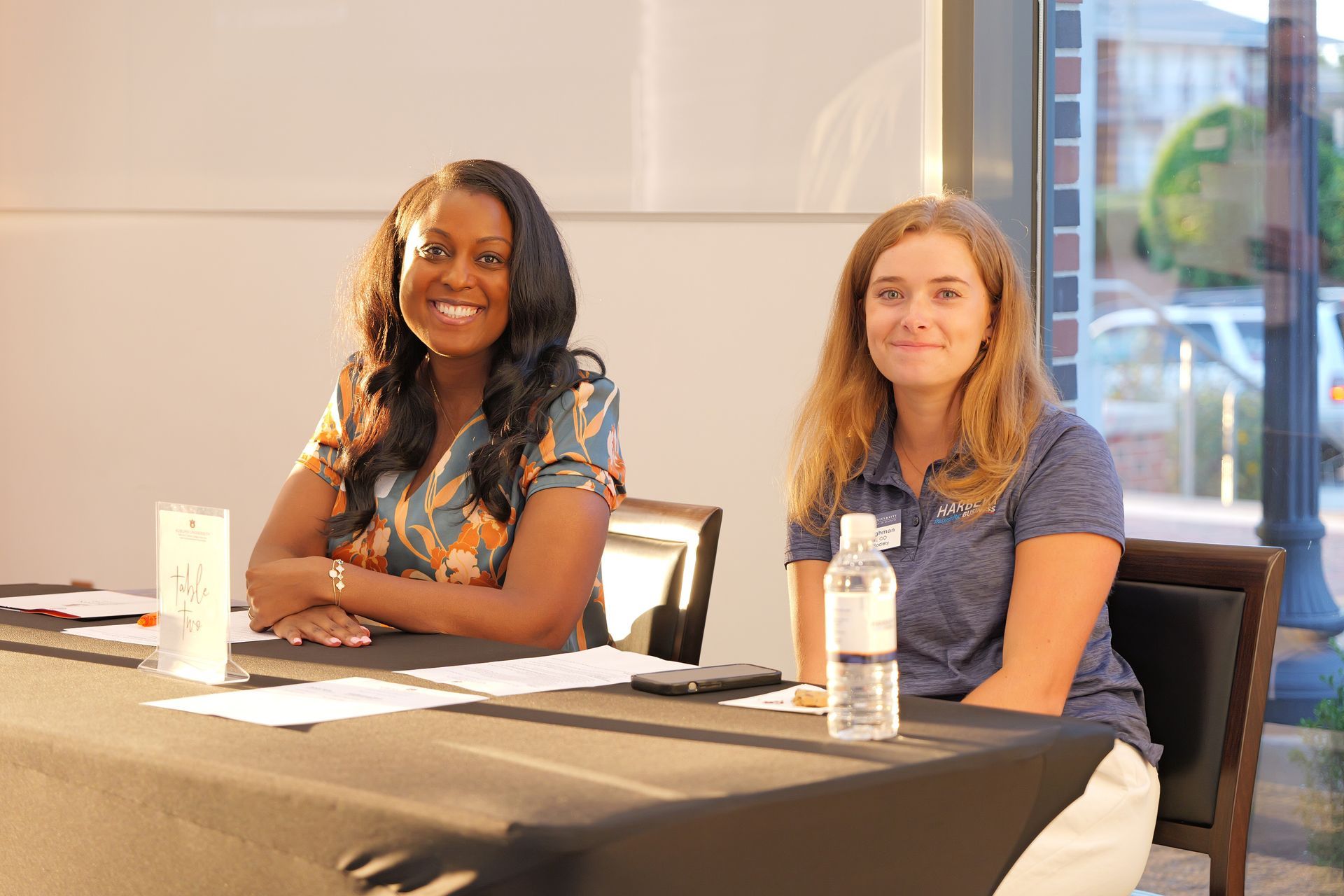 Two women sit at a table indoors, smiling at the camera.