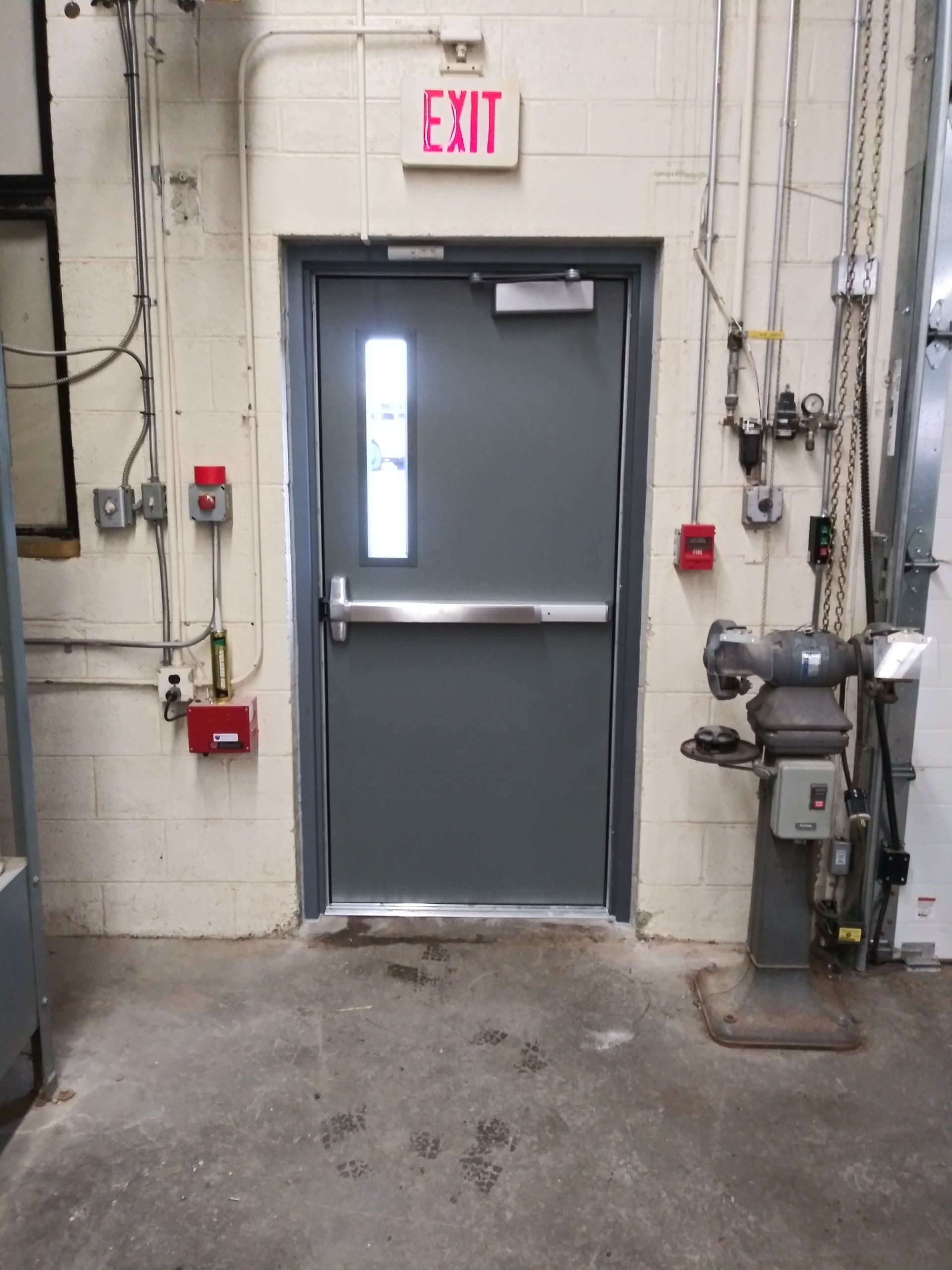 Gray industrial exit door with red EXIT sign in a utility room, surrounded by electrical equipment and pipes