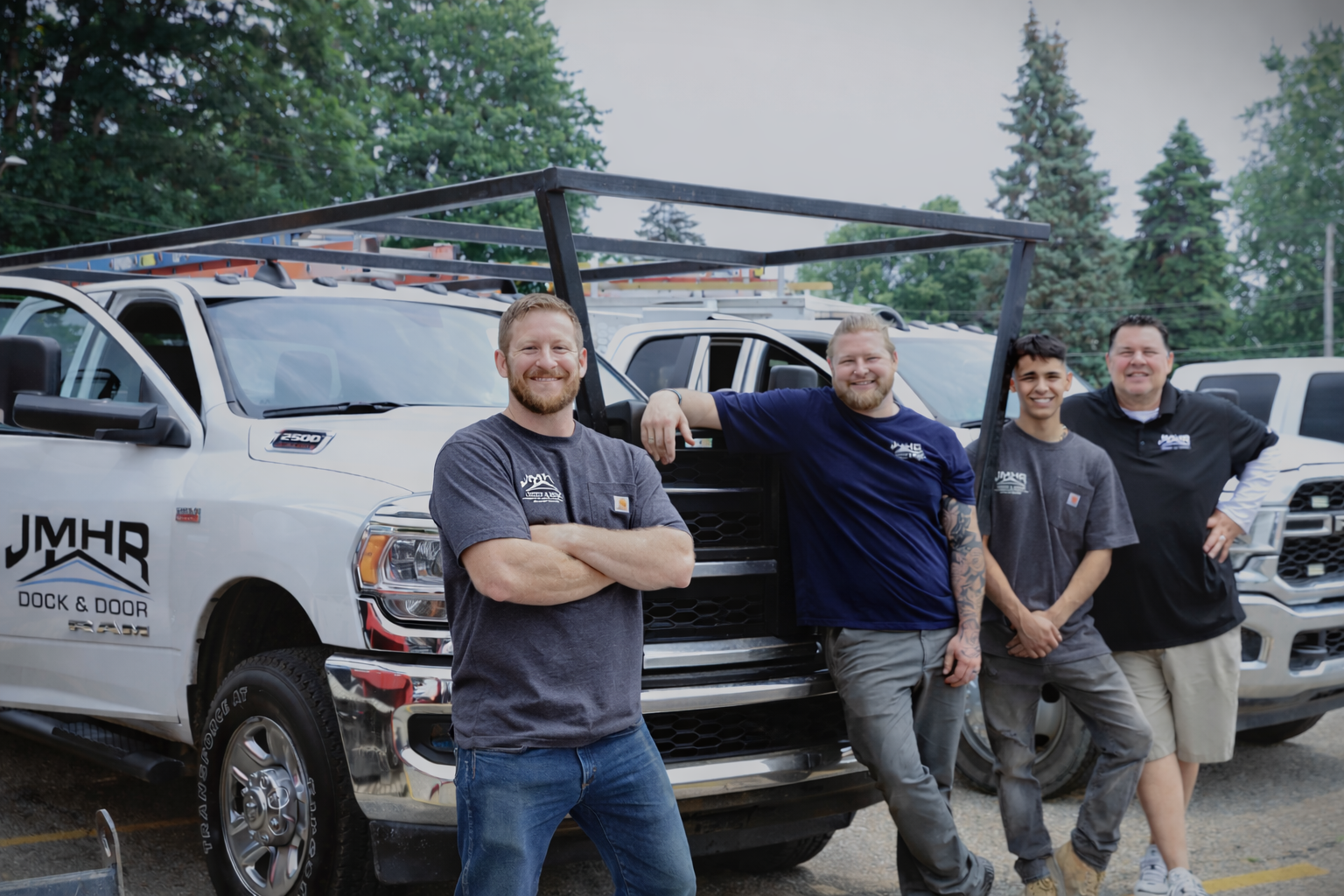 Four men stand near white work trucks with a rack. One man has arms crossed, two are leaning, all are smiling.