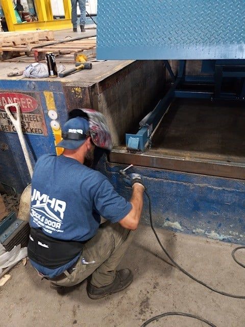 Worker kneeling beside a blue truck, inspecting a compartment with a flashlight and tool on a roadside.