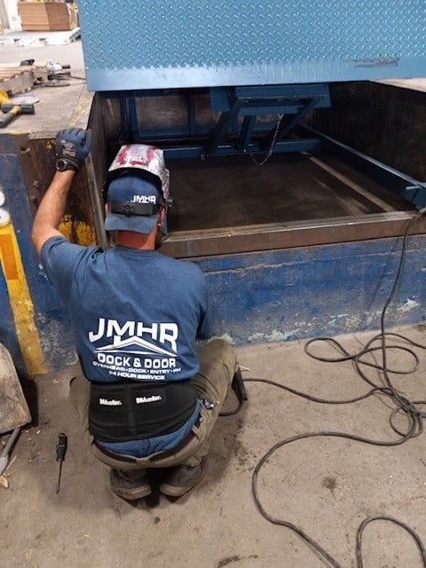 Worker kneels beside a large blue industrial machine in a workshop, handling cables and equipment.