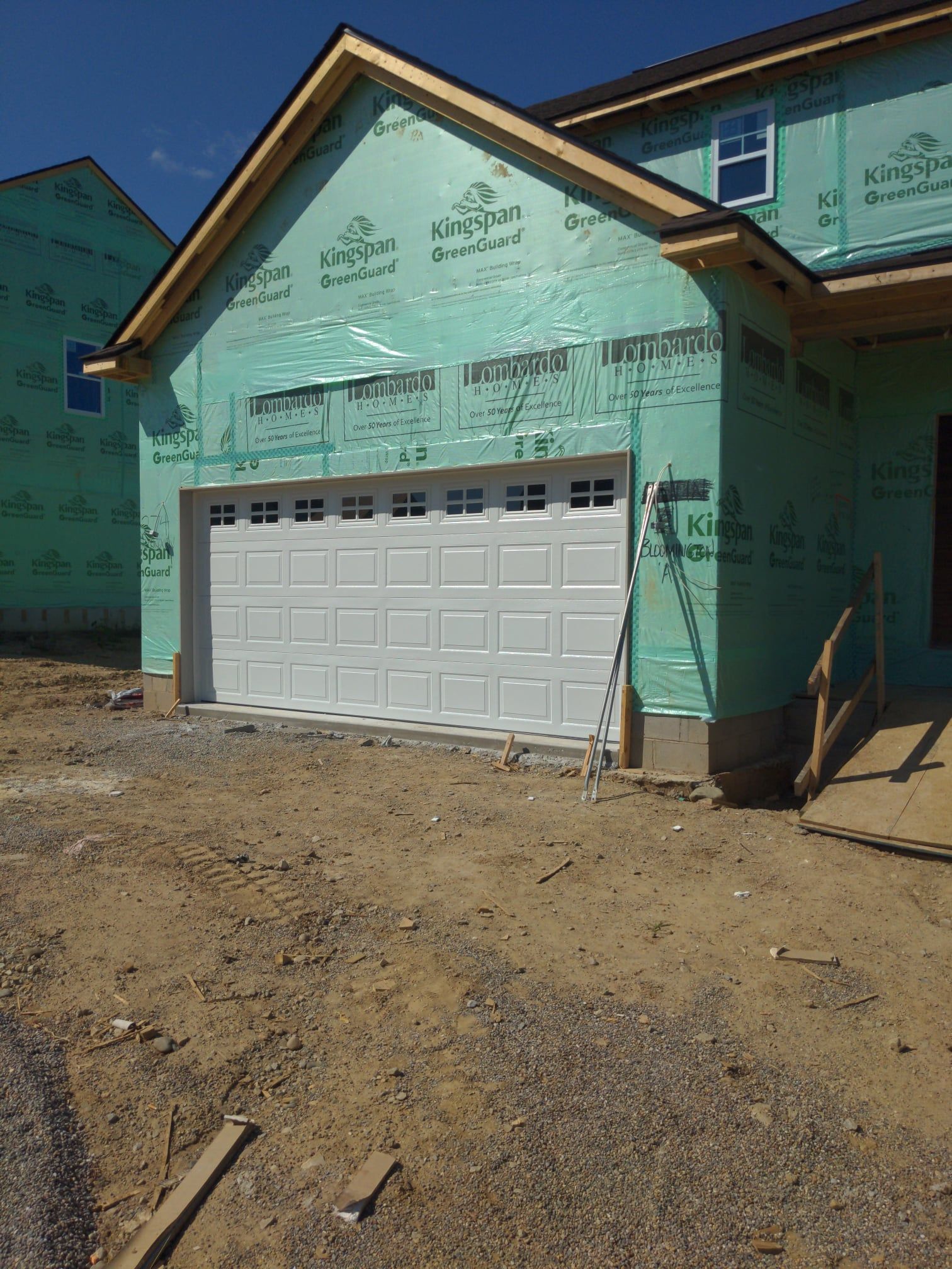 Partially finished house with green exterior wrap and white garage door on a dirt lot