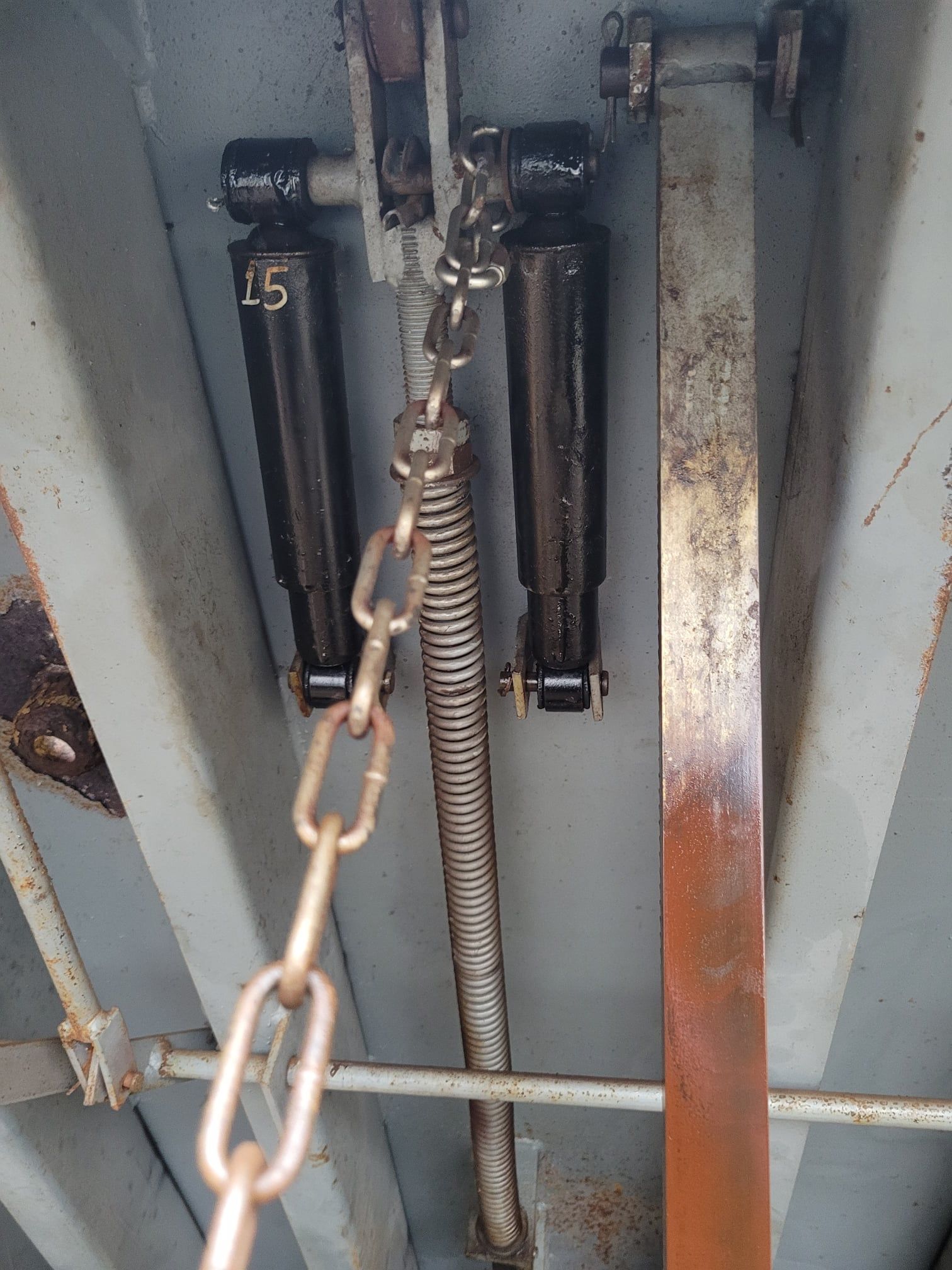 Close-up of a metal spring and black components inside a gray industrial cabinet with a rusty panel.