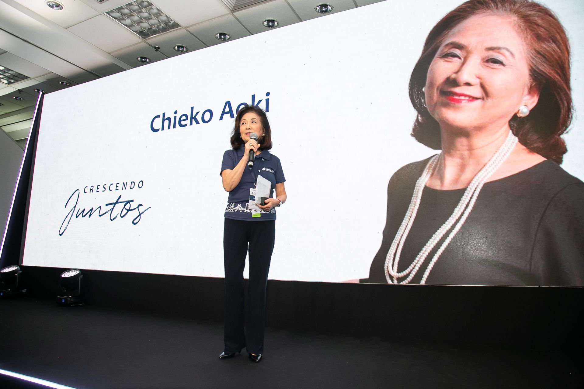 Uma mulher está parada em frente a uma tela grande que diz chieko aoki