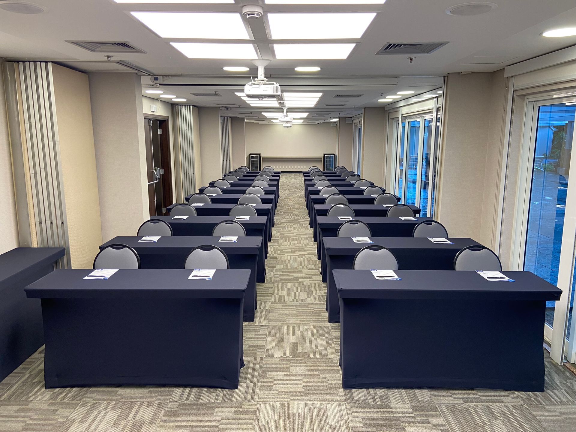 Uma sala longa com fileiras de mesas e cadeiras e um projetor.