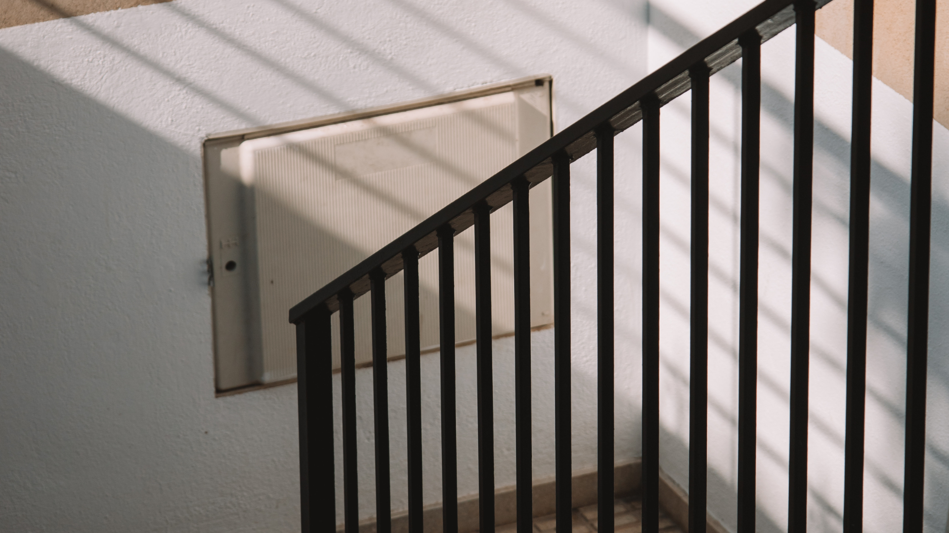 Black stair railing next to white wall with rectangular panel. Sunlight casts shadows.