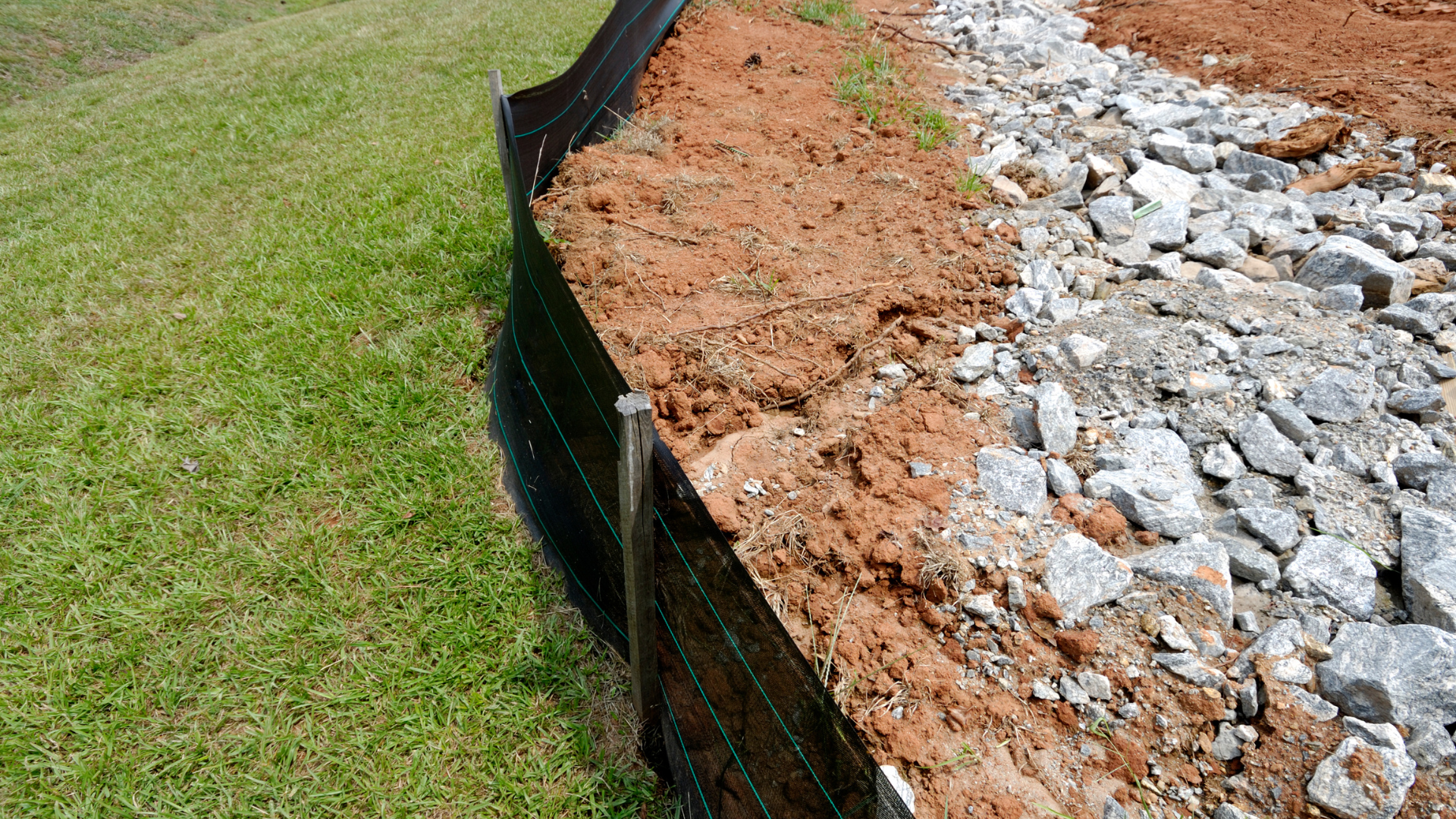 Black silt fence separating green grass, dirt, and rocks.