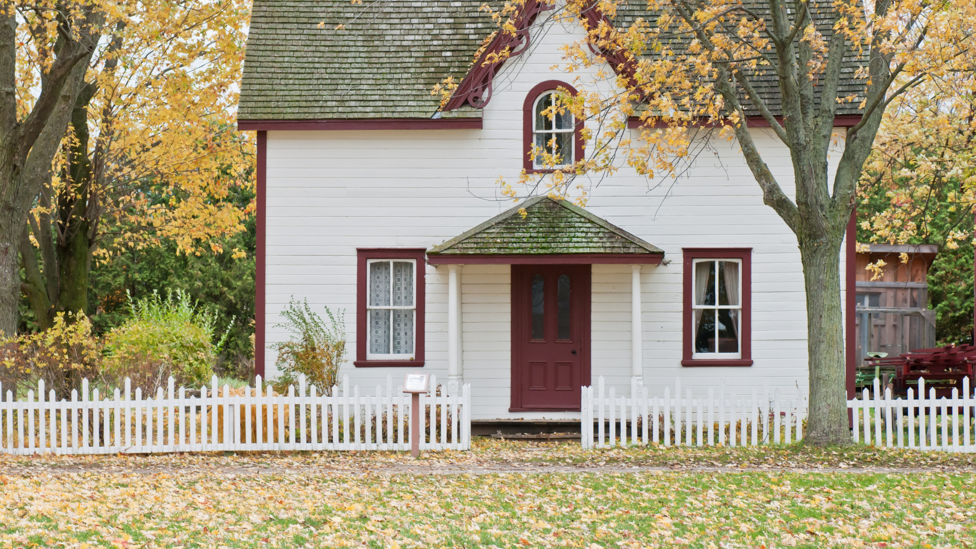 White cottage with red trim and picket fence, set among fall foliage.