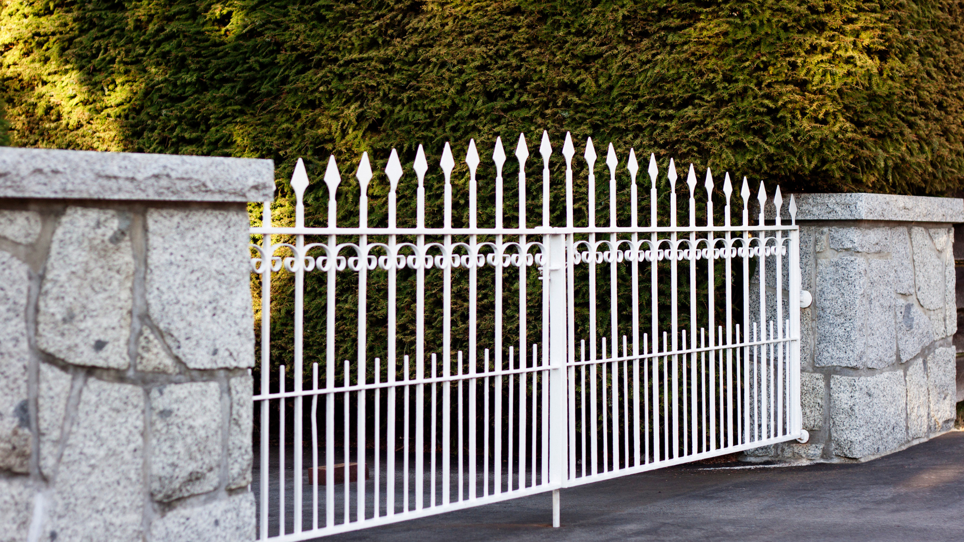 White wrought iron gate and stone wall in front of green foliage.