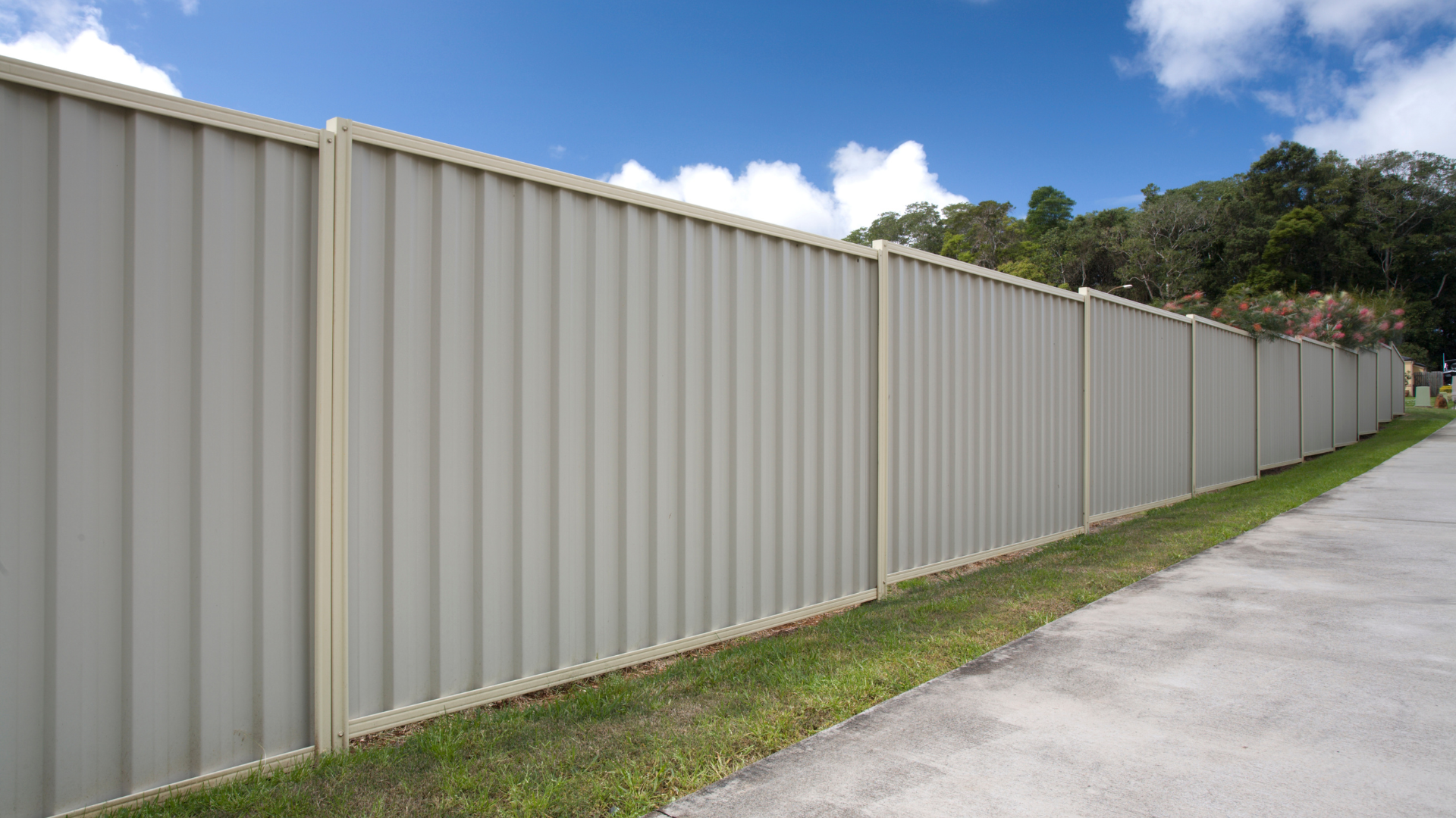 Beige corrugated metal privacy fence bordering a grassy area and asphalt road under a blue sky.