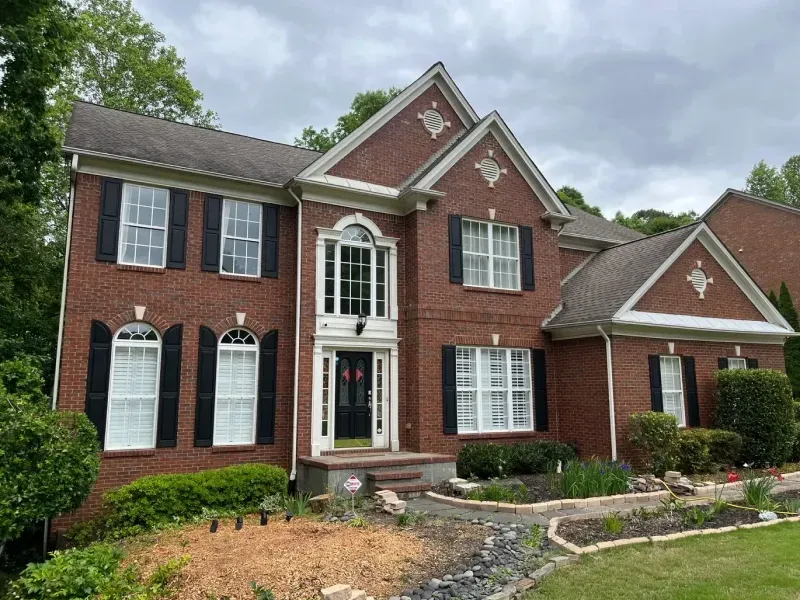 Two-story brick house with black shutters, white trim, and a front garden on a cloudy day.
