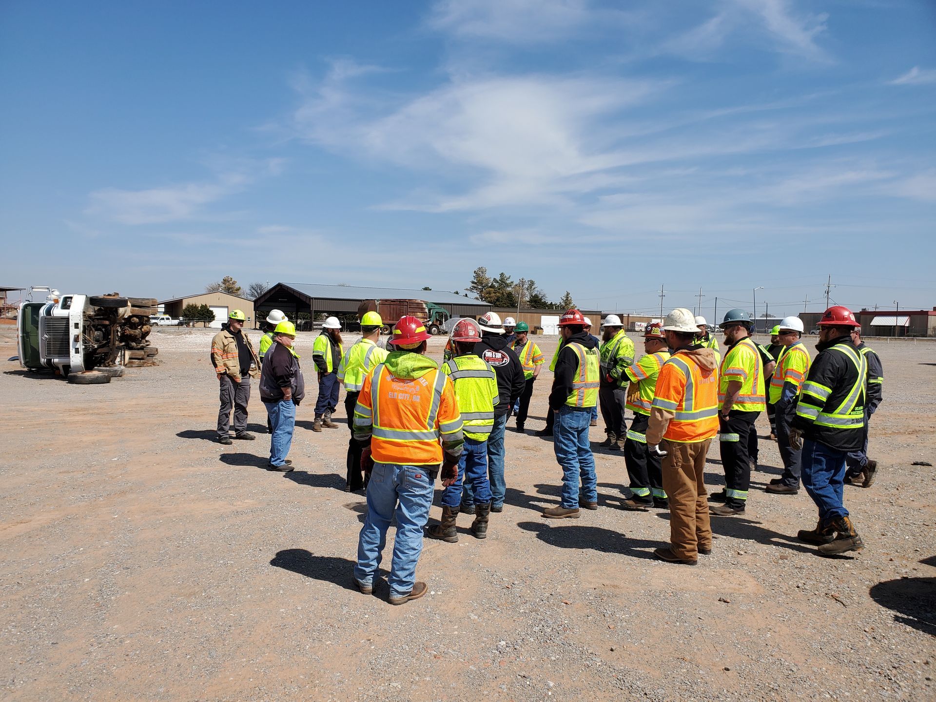 Construction workers in hard hats and orange vests gathered on a gravel site under a blue sky.