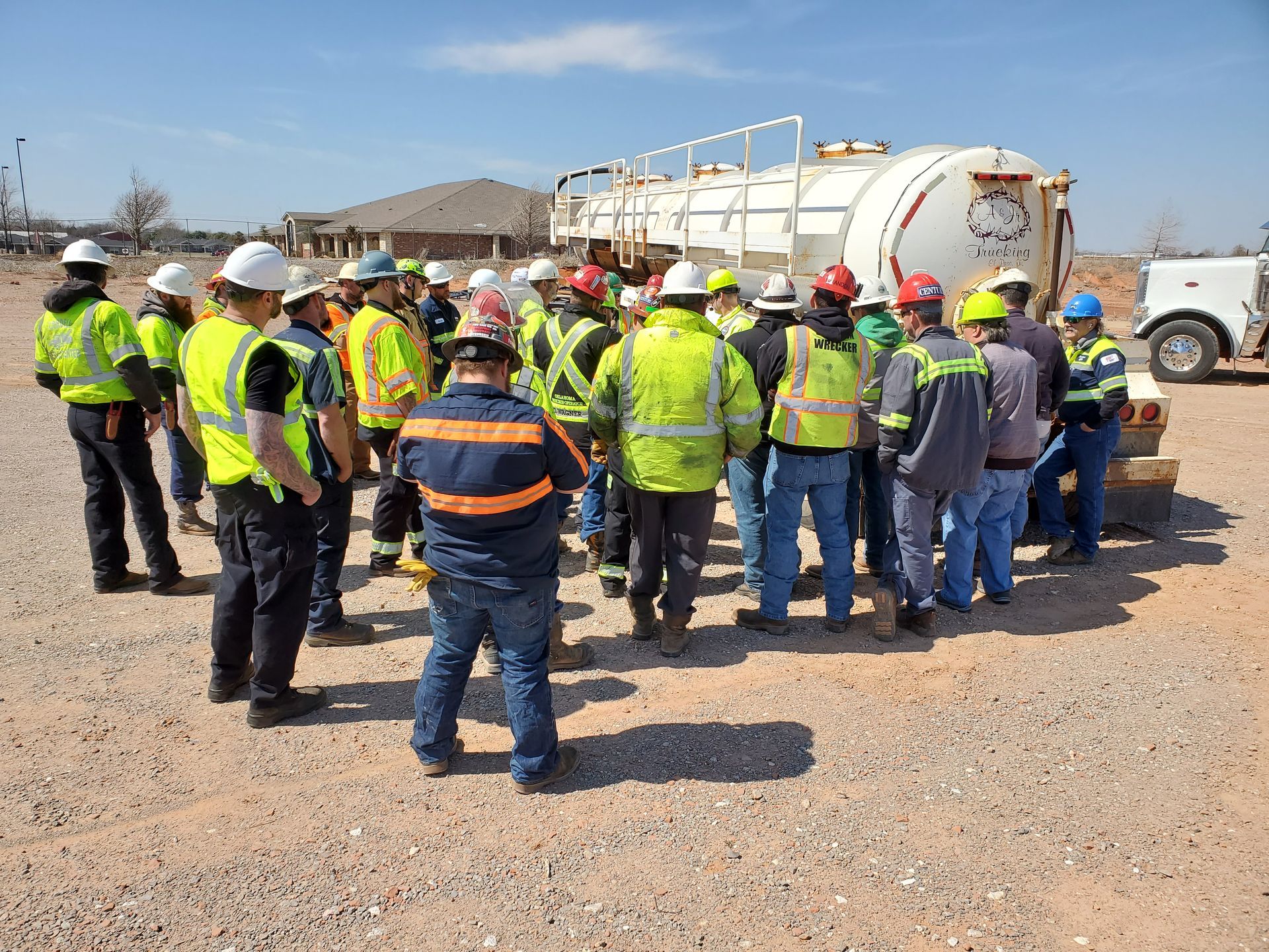 Workers in safety vests and hard hats gather around a large tanker truck on a gravel site.