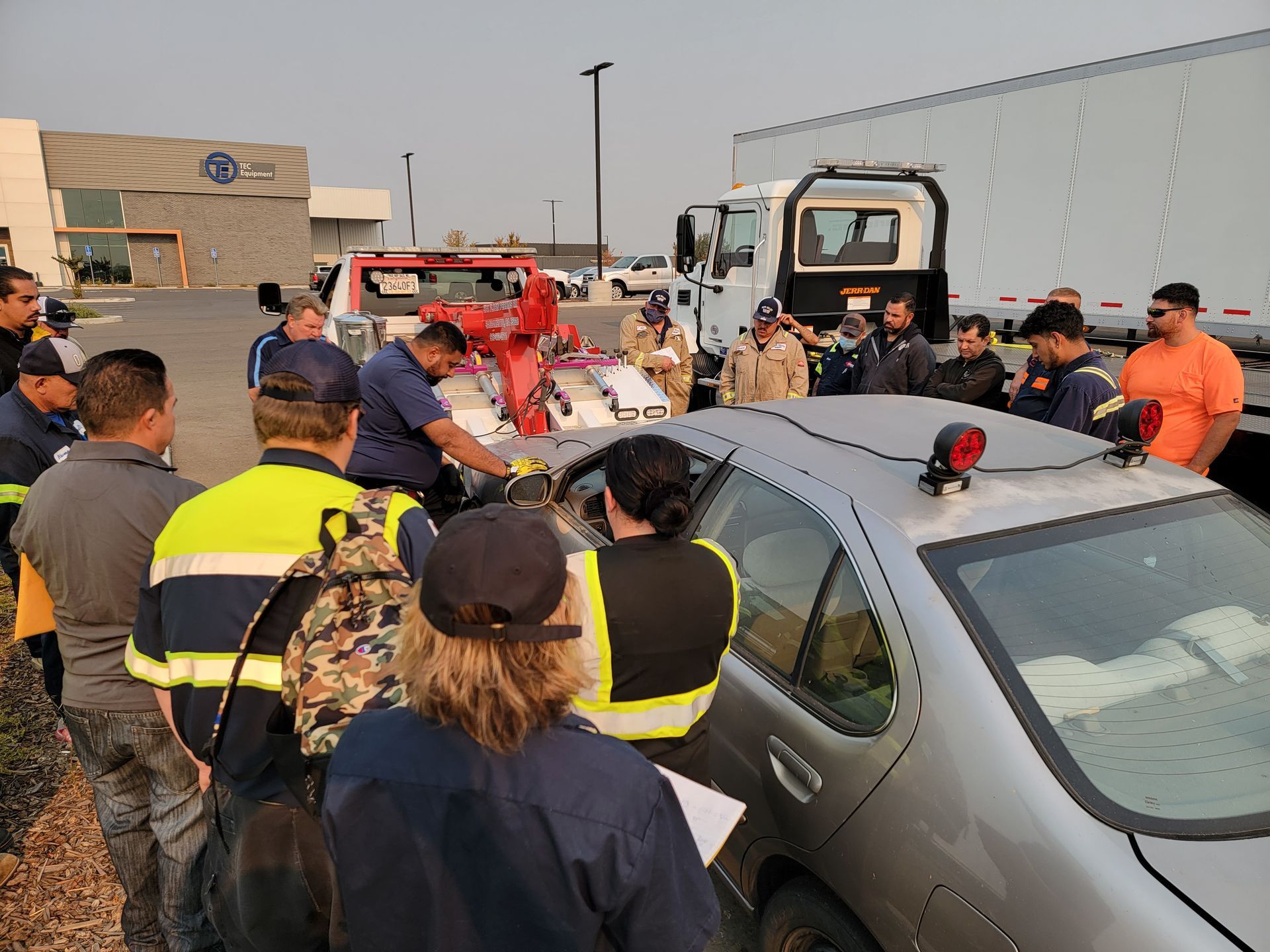 Emergency responders crowd around a gray car at a crash scene beside a truck and trailers.