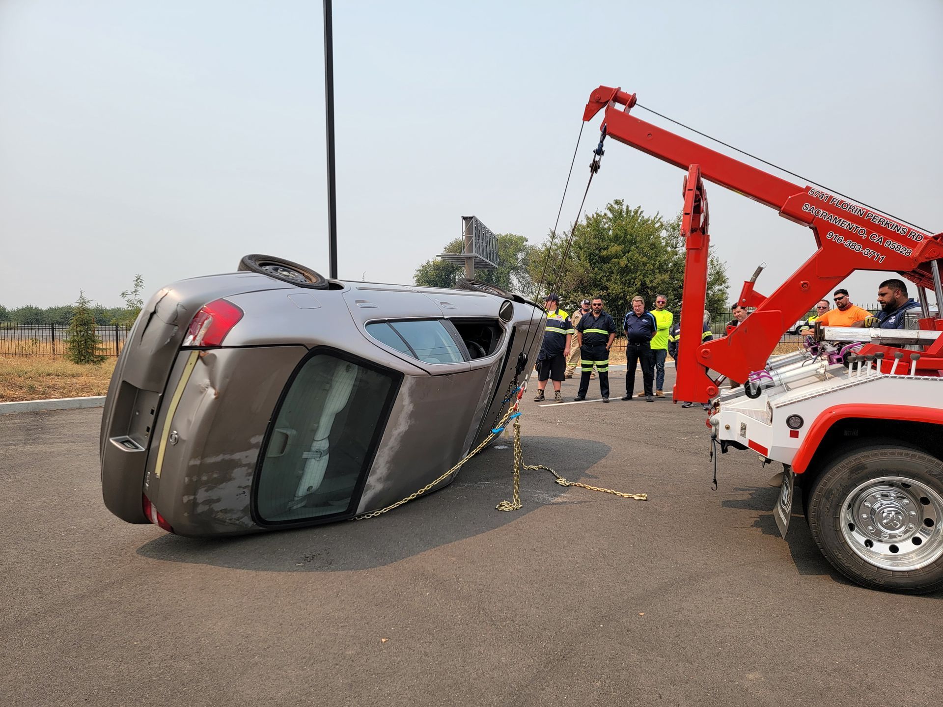 Overturned gray car on roadside beside red tow truck, with people gathered nearby.