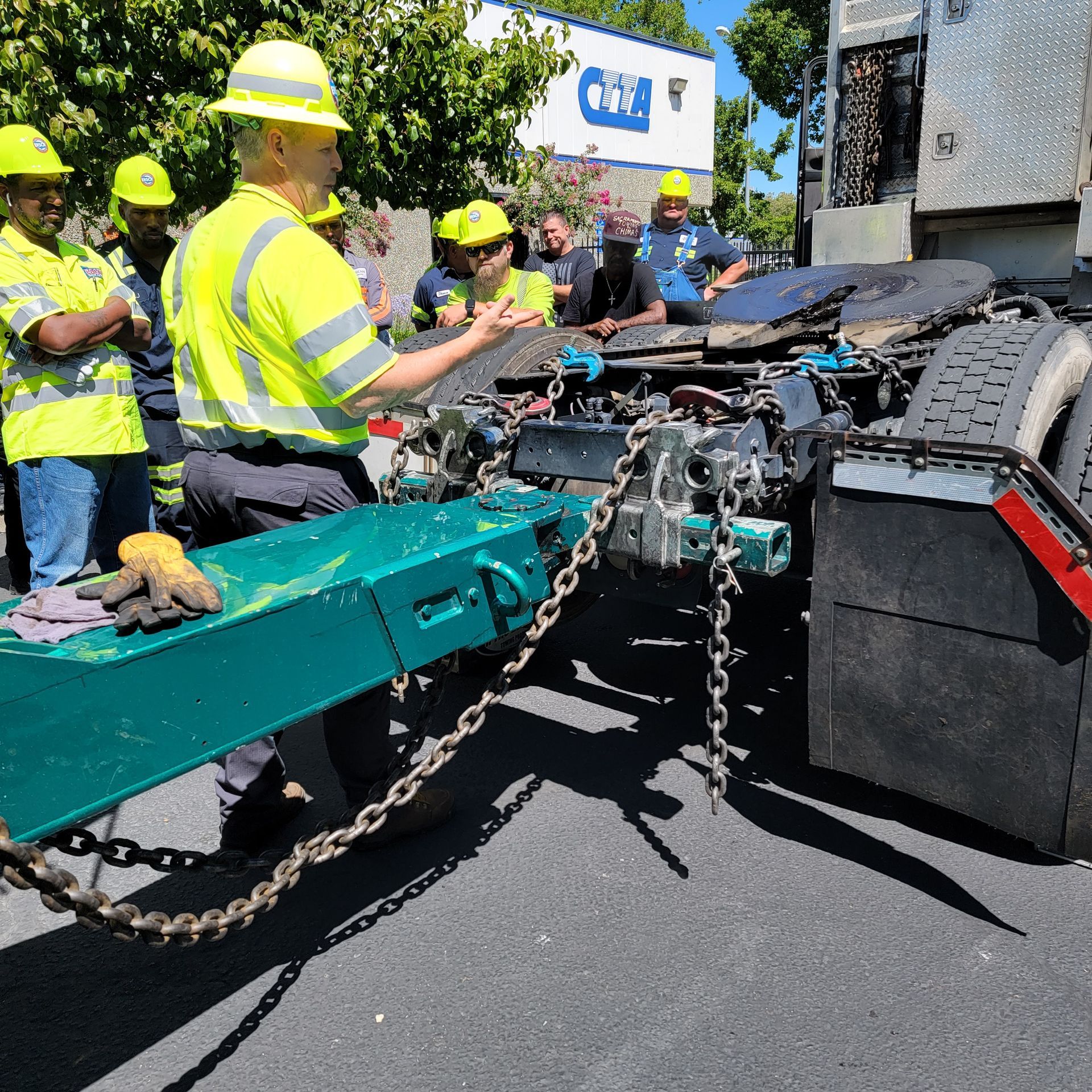 Workers in yellow hard hats inspect a tipped teal flatbed truck on a roadside.