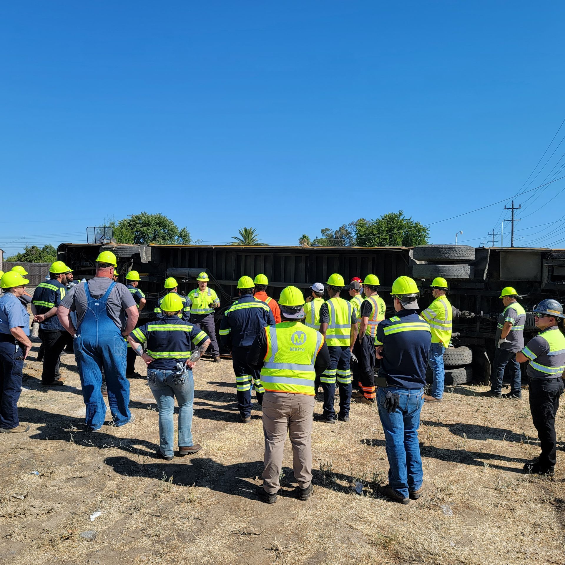 Construction crew in yellow hard hats gathered outdoors beside a large overturned vehicle.