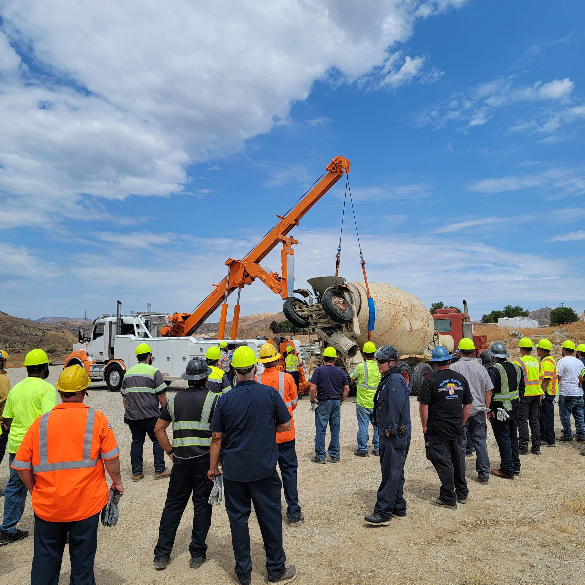 Workers in hard hats watch a crane lift a large concrete pipe at a construction site under a blue sky