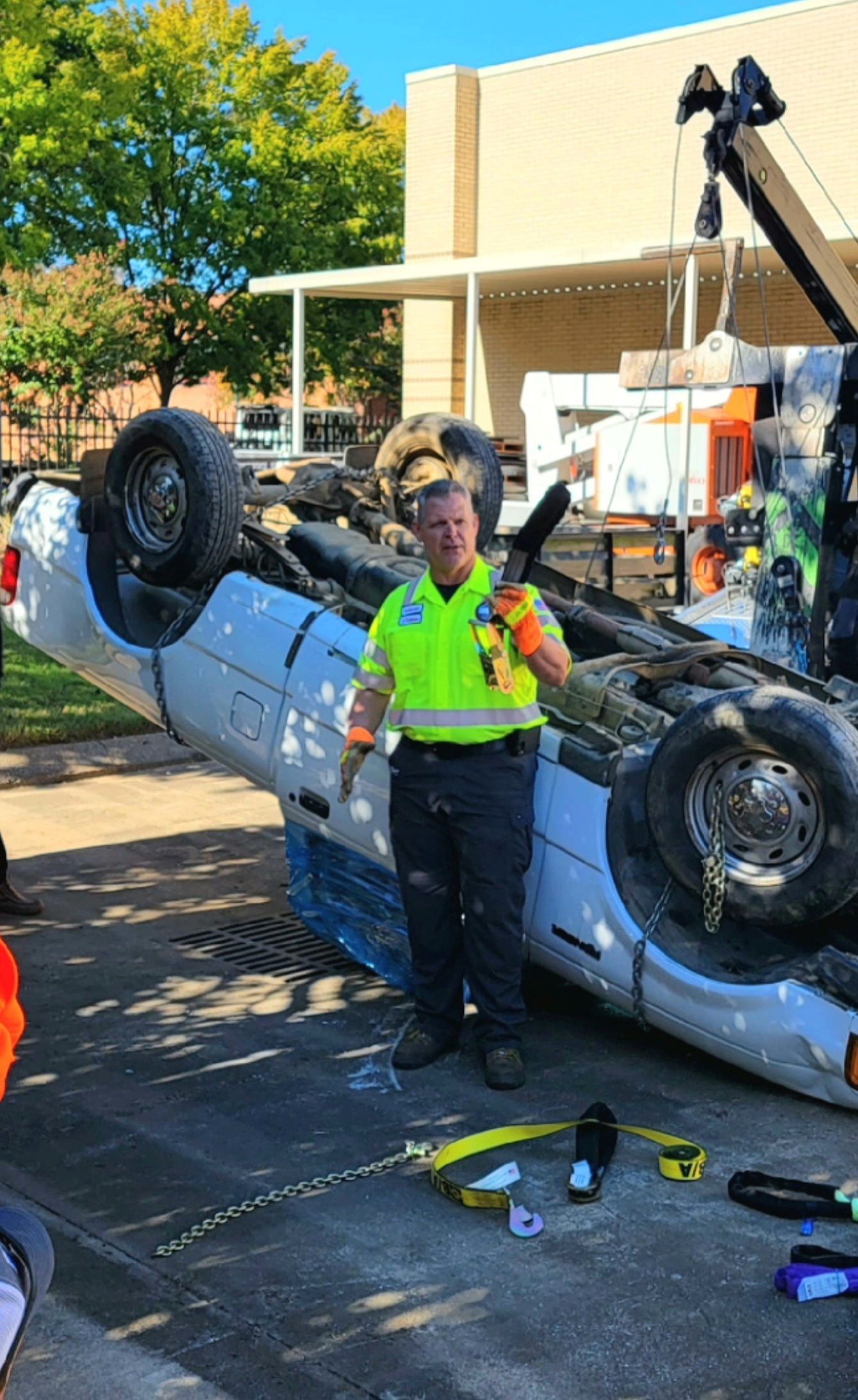 Emergency responder stands beside an overturned white car in a sunny street scene.