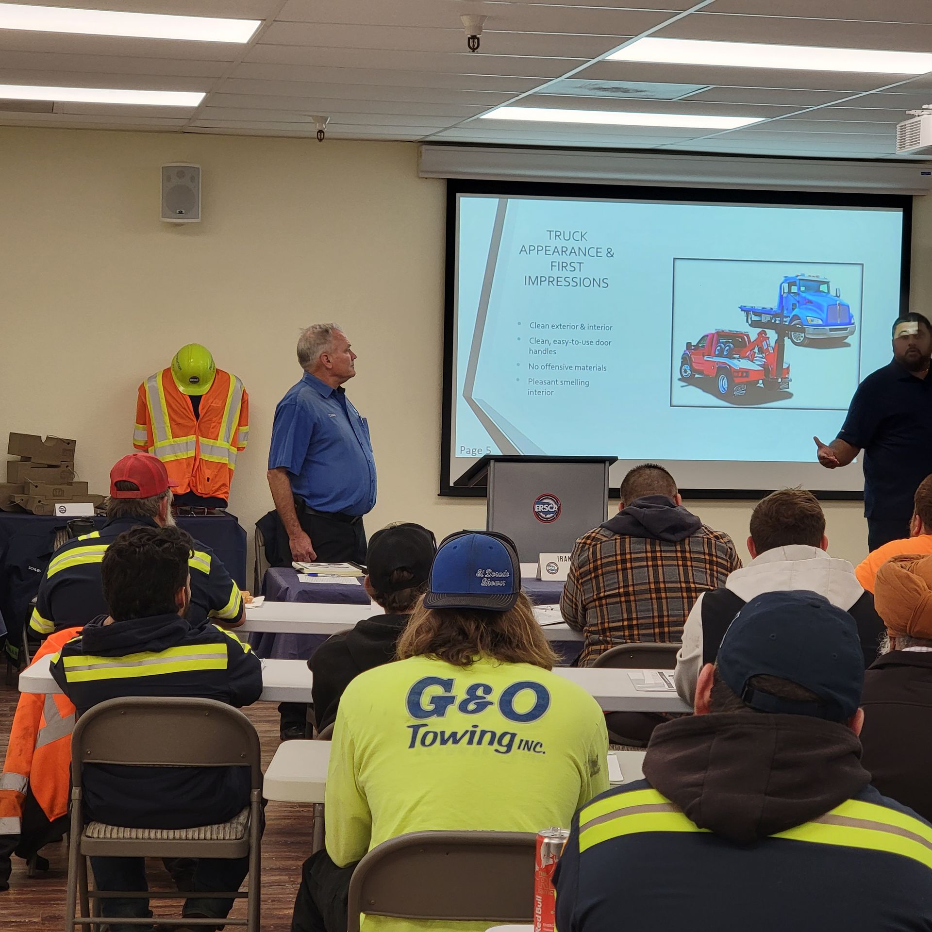 Towing safety presentation in a classroom, with an instructor at a projector and workers seated in reflective gear