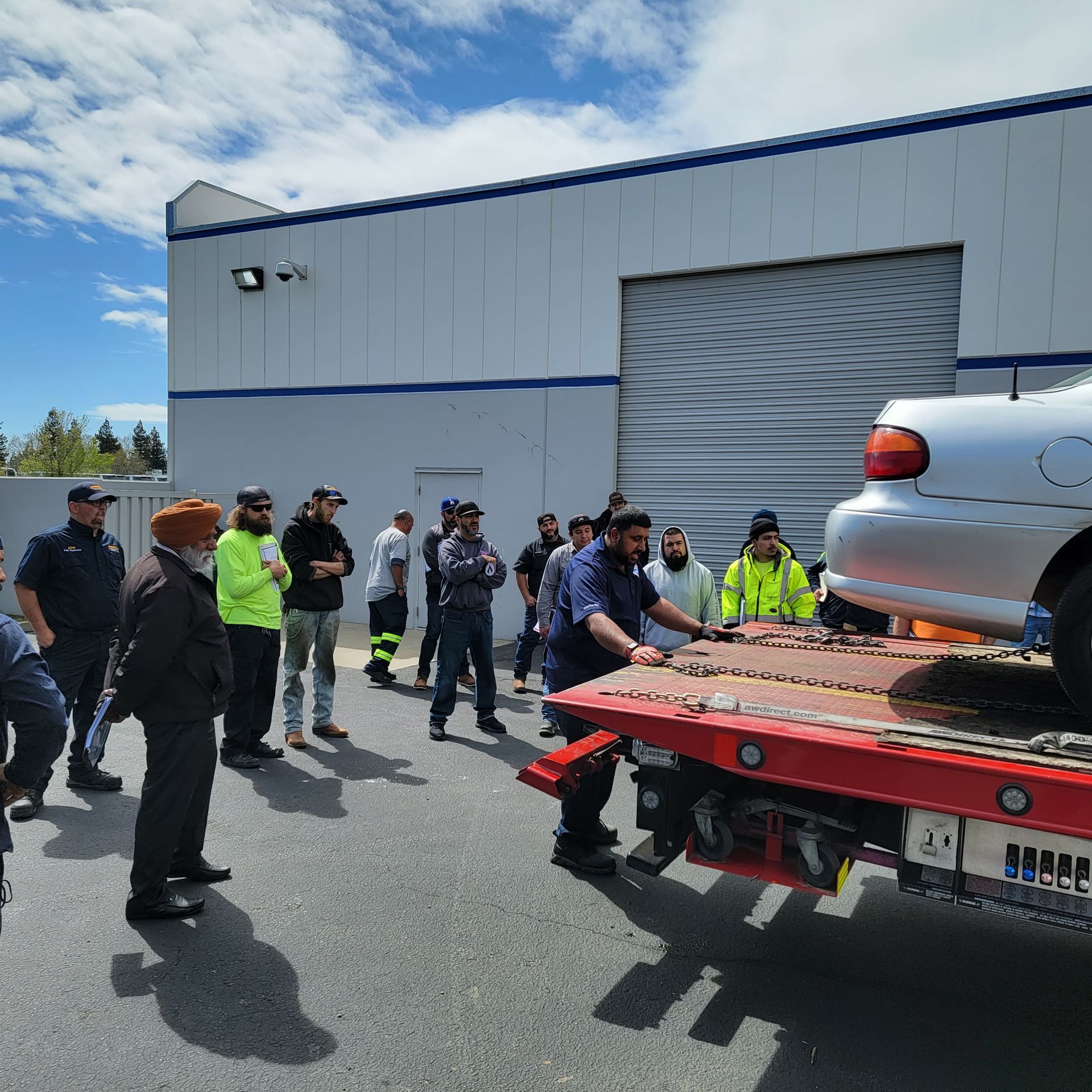 Workers and officers watch a car loaded on a flatbed tow truck outside an industrial building.