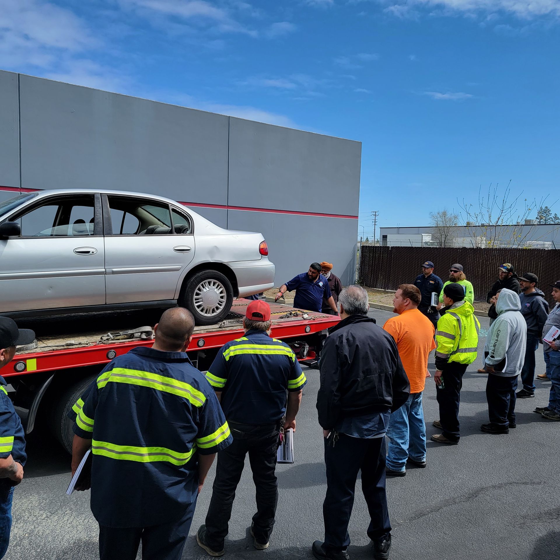 Tow truck loading a silver sedan while workers in safety gear watch in a parking lot under a blue sky