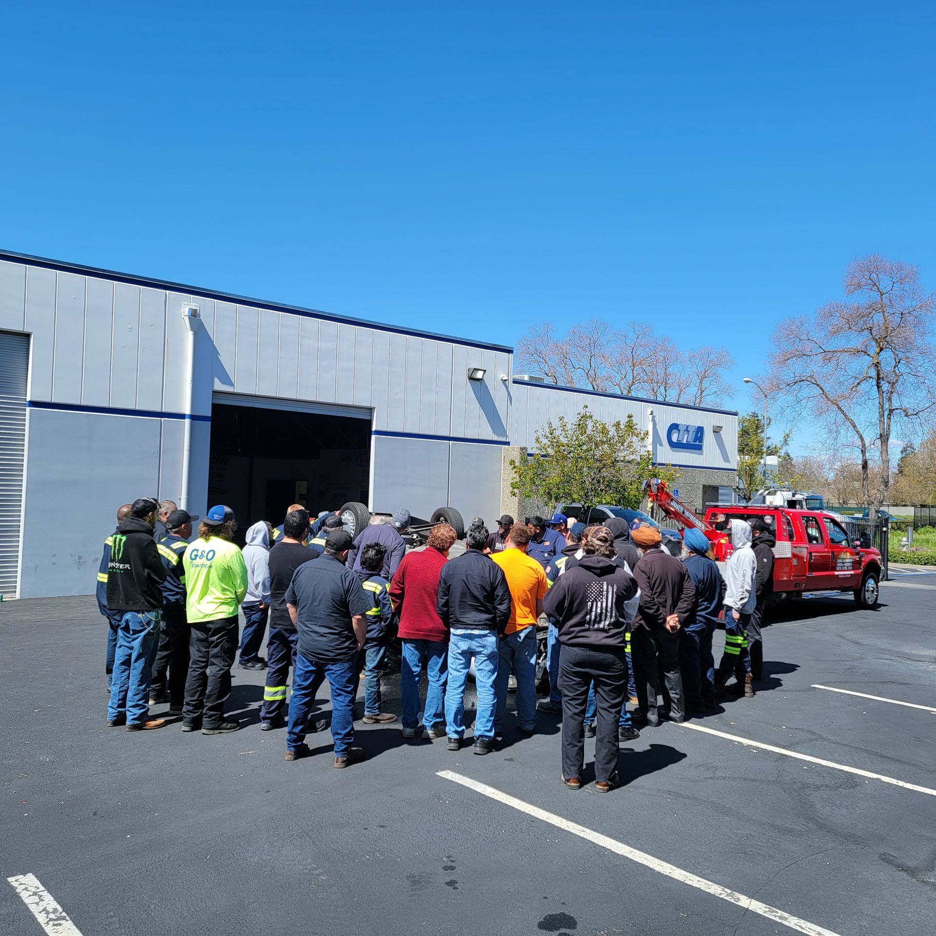 Group of people gathered outside a gray industrial building beside parked vehicles on a sunny day