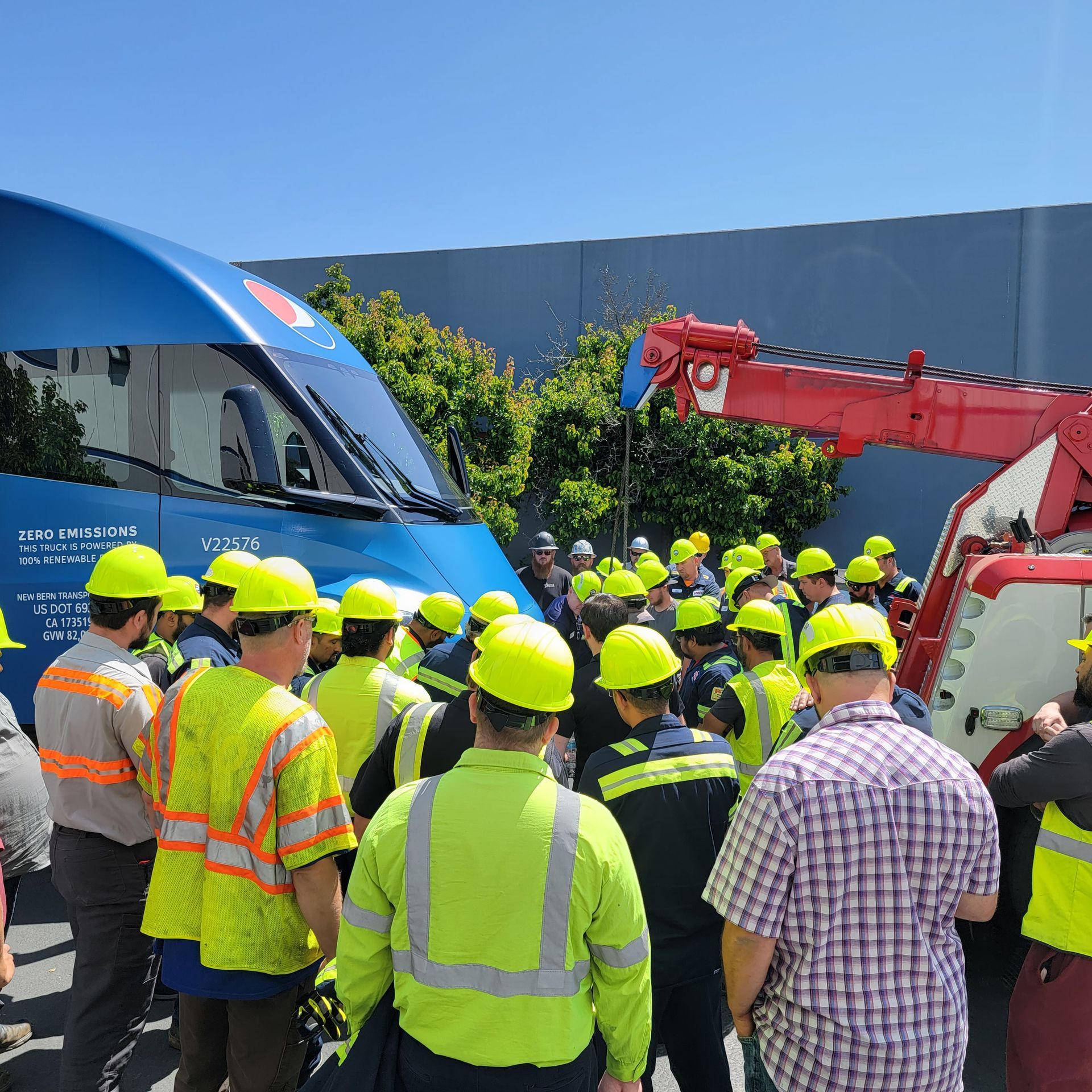Workers in yellow hard hats and safety vests gather beside a blue train and red crane outdoors.