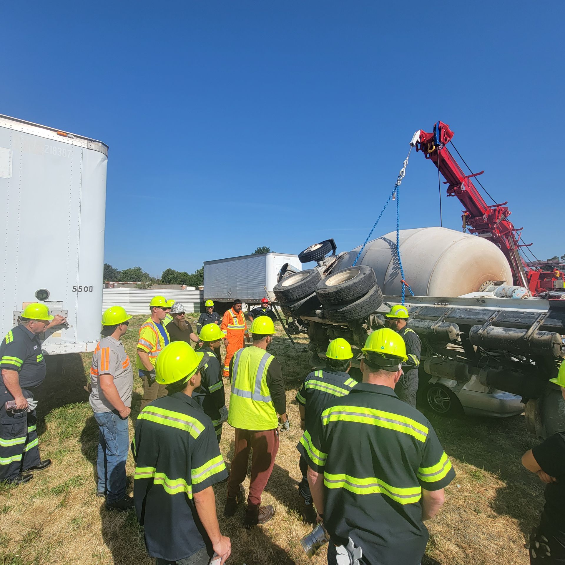 Firefighters in yellow helmets gather around an overturned tanker truck at a roadside crash scene.