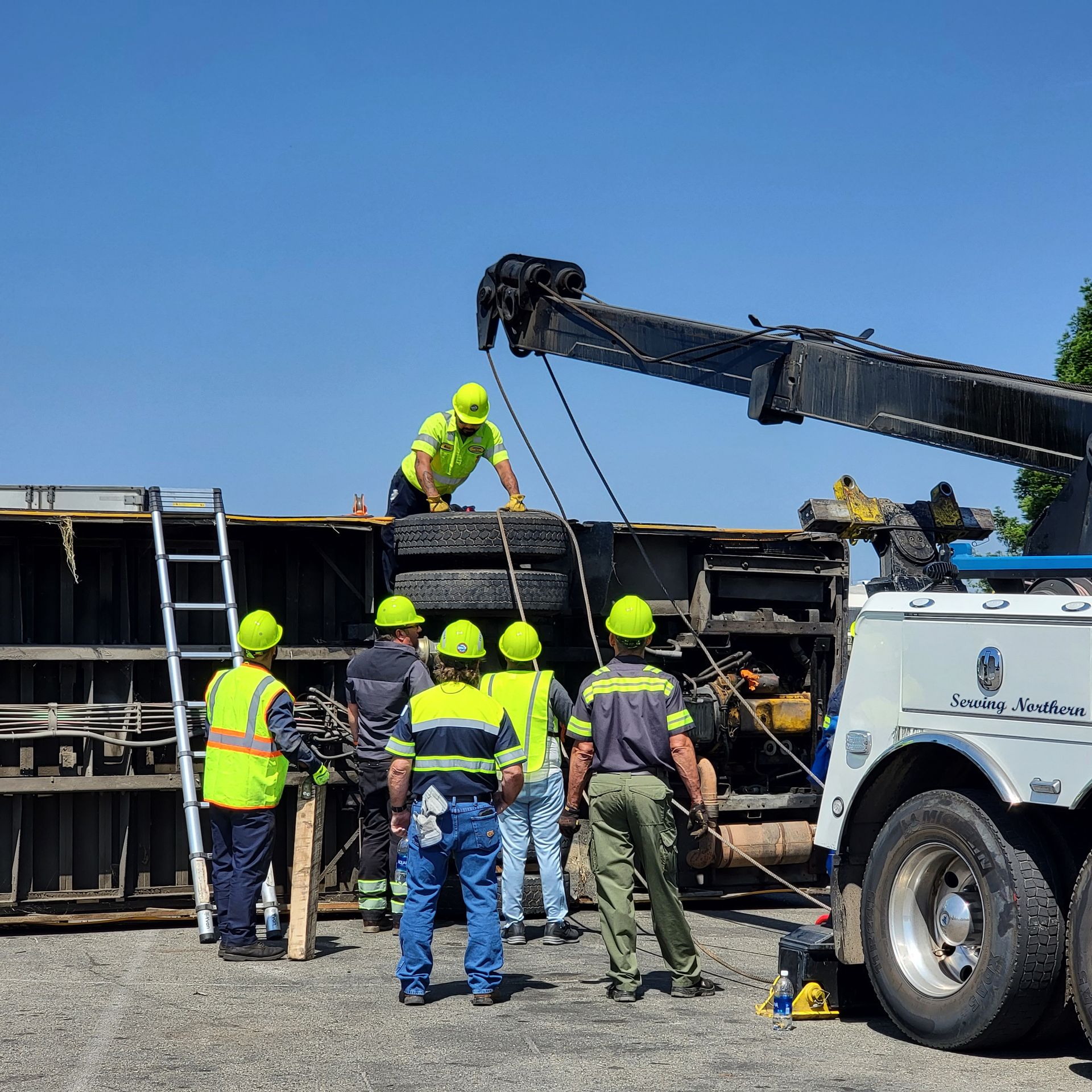 Workers in neon hard hats inspect an overturned truck beside a tow crane on a sunny roadside.