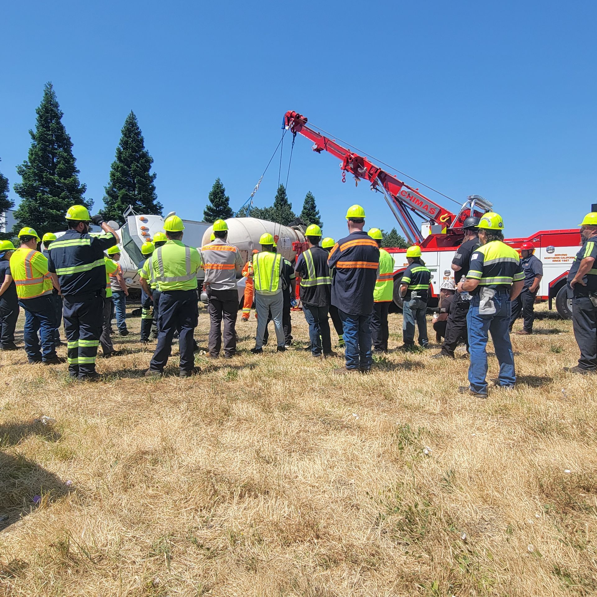Firefighters and workers gather in a dry field near a red crane under a clear blue sky.