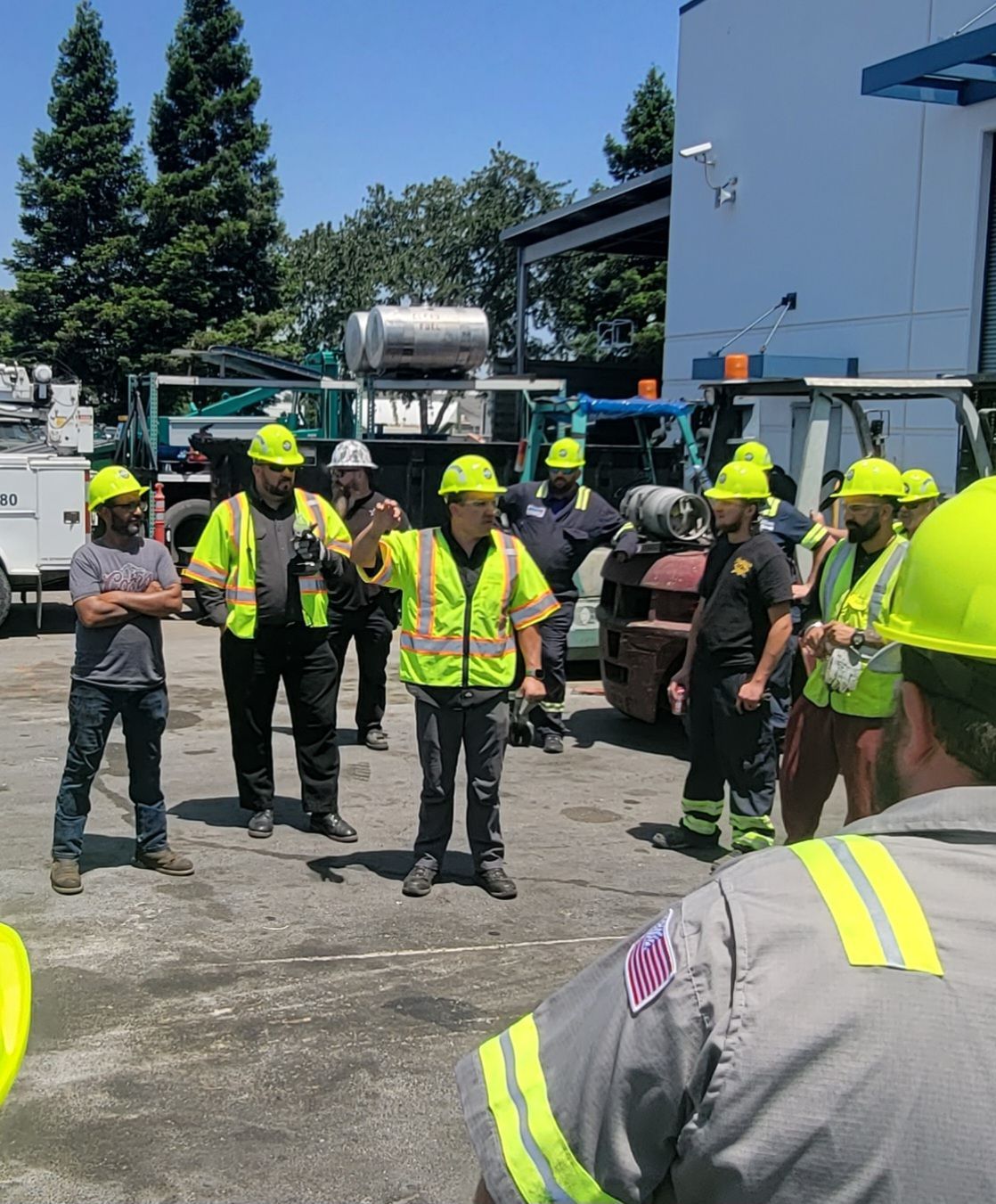 Workers in hard hats and safety vests gathered in a construction yard, facing a speaker.