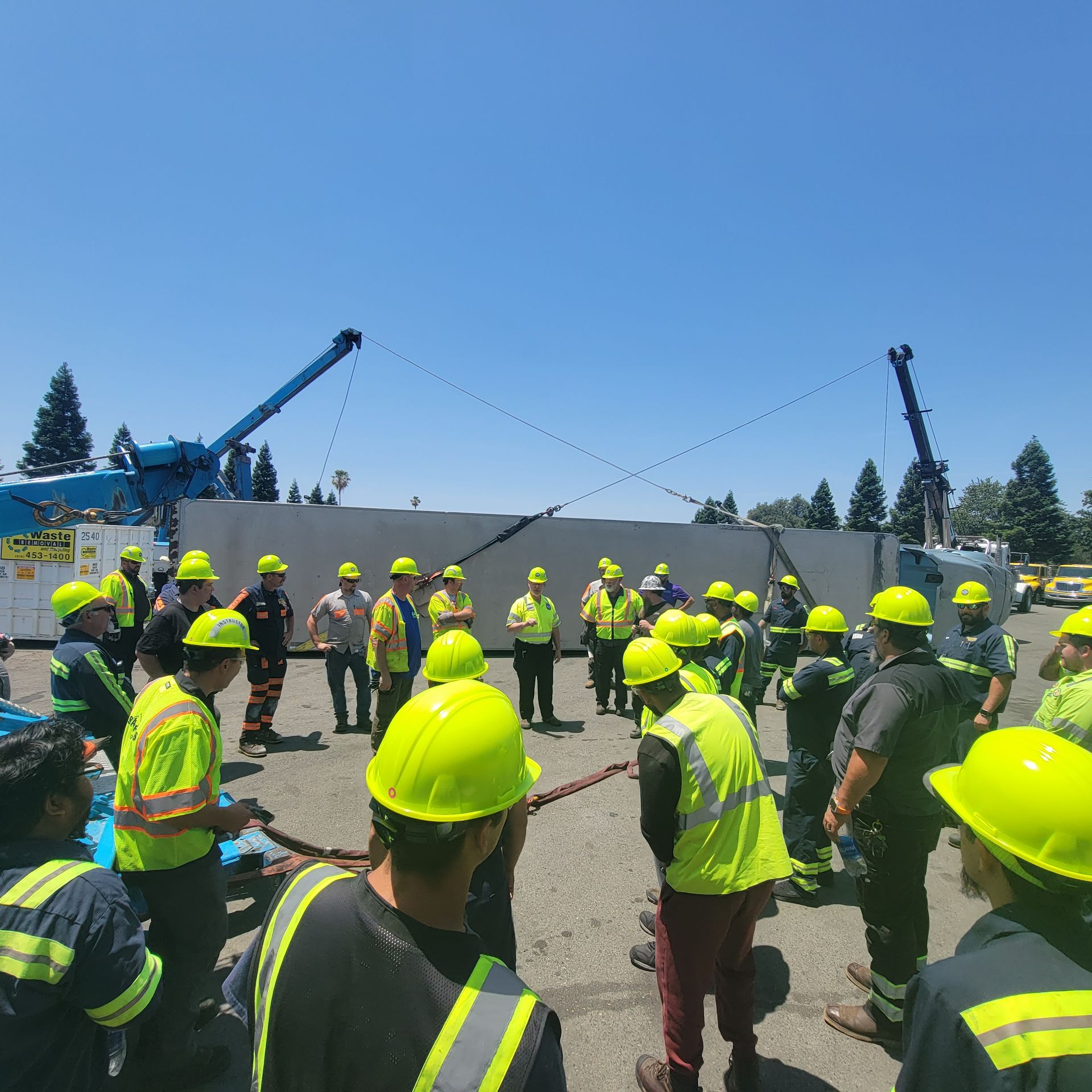 Construction crew in neon safety vests and hard hats gathered around a large concrete wall and cranes outdoors
