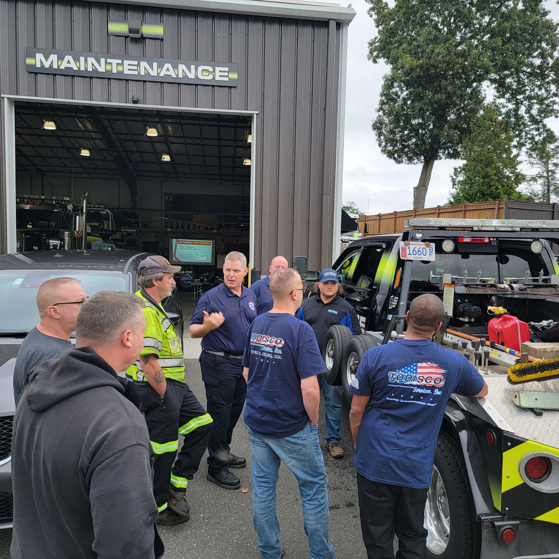 Workers and firefighters gather outside a maintenance garage beside a tow truck and equipment.