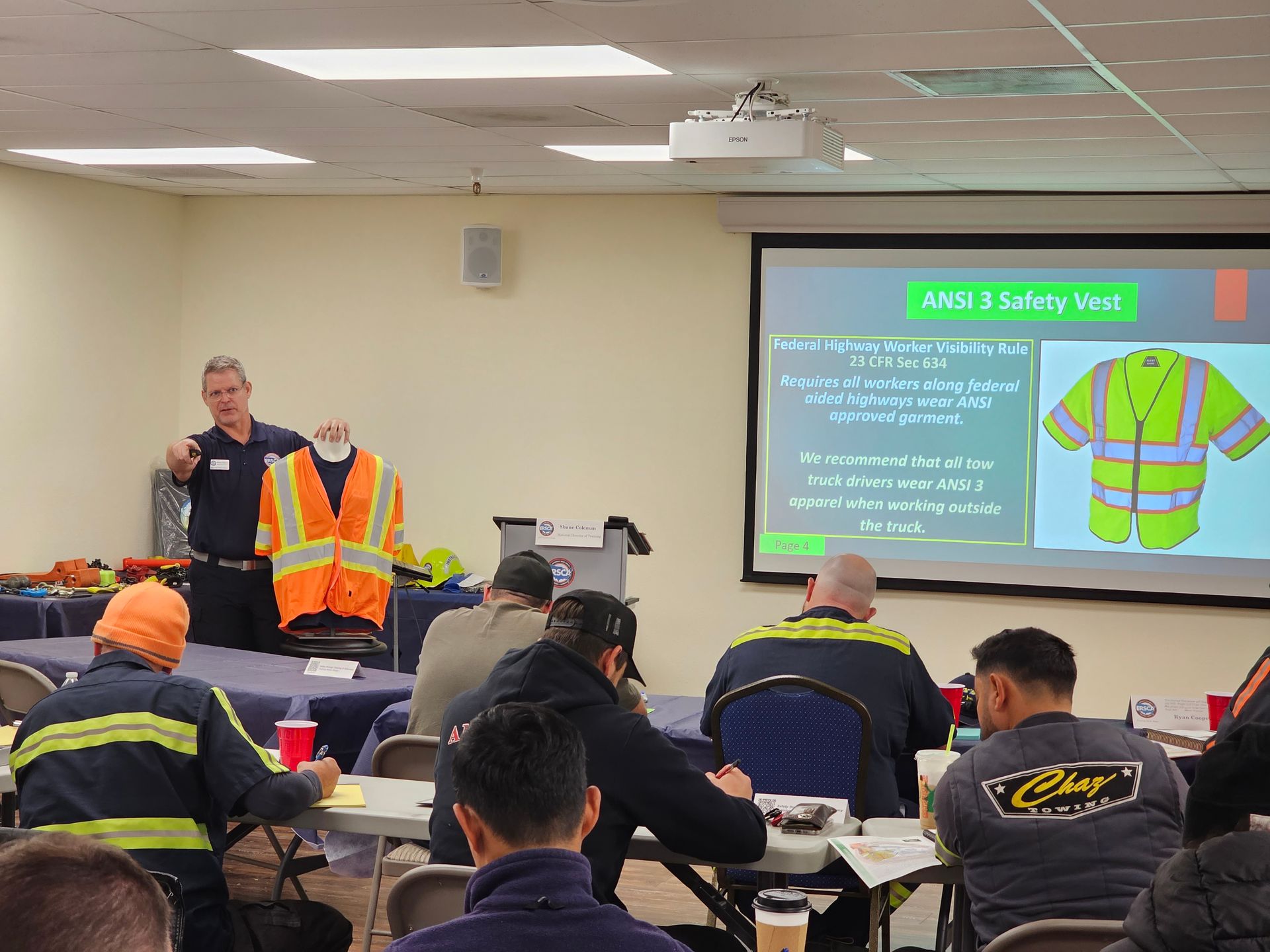 Trainer presents safety vest at classroom session with seated workers and projected slide on the screen