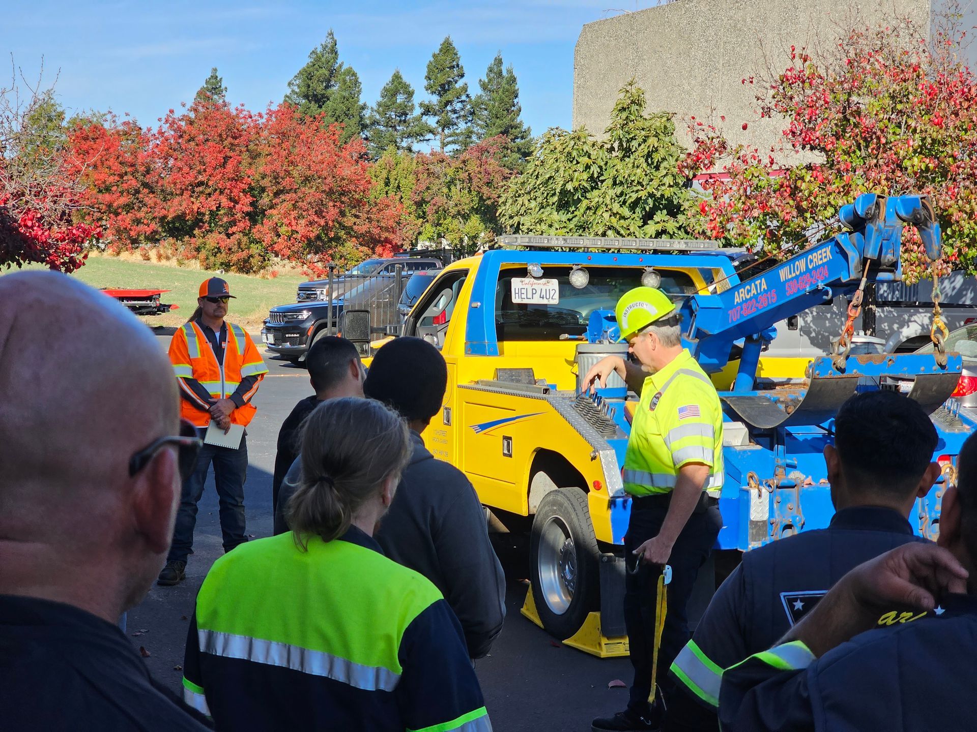 Crowd watching workers in safety gear beside a yellow tow truck at a roadside scene