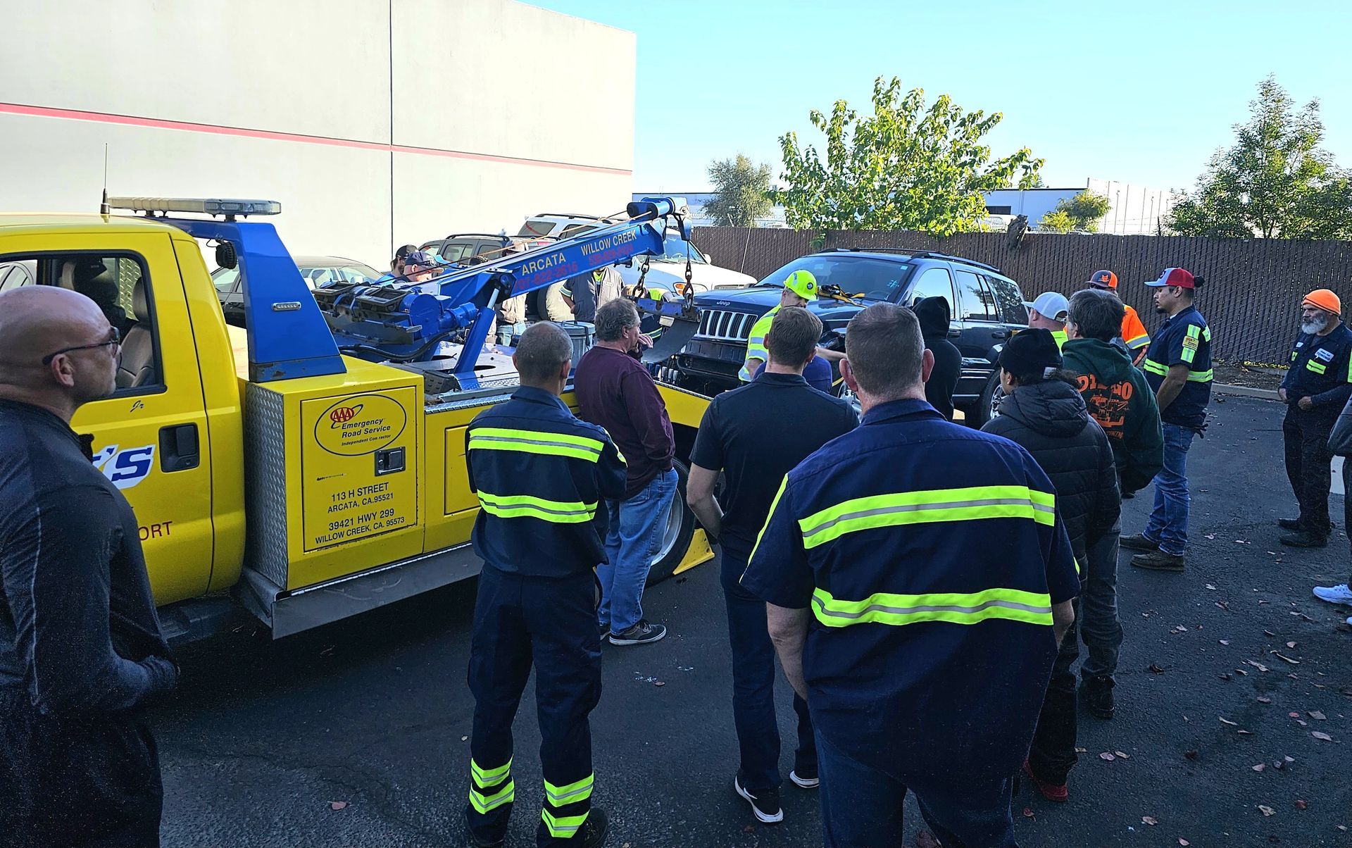 Workers and emergency responders gather beside a yellow tow truck in a lot, some wearing reflective vests.