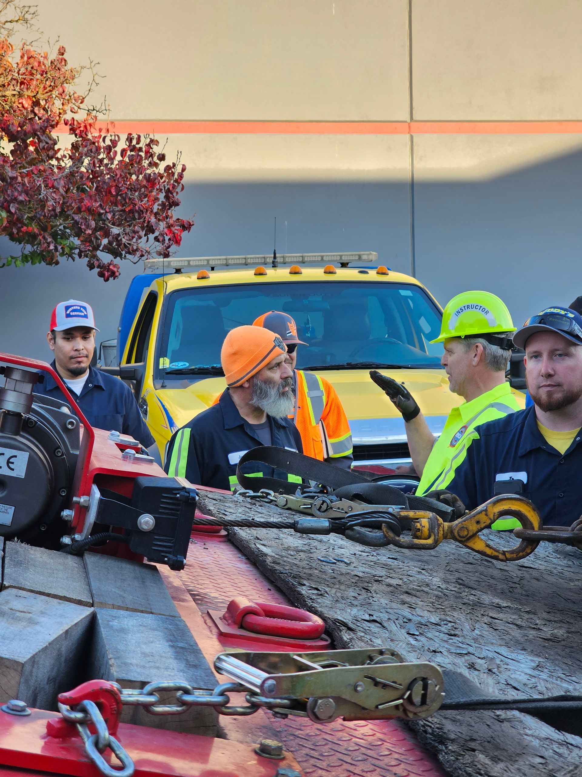 Workers inspect a damaged tow truck rig with chains and straps at a roadside work site