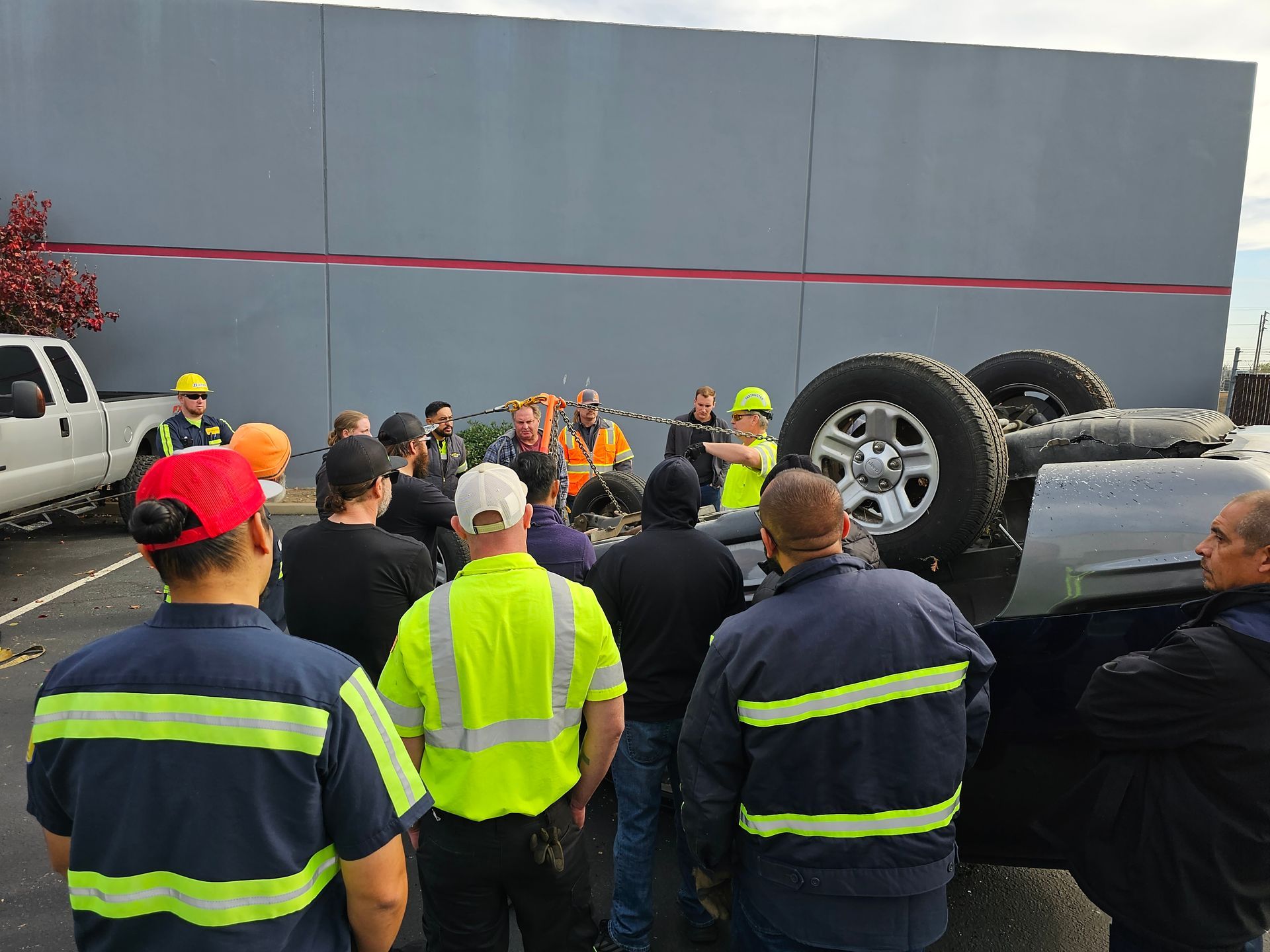 Workers and bystanders gather near an overturned SUV on its side at an outdoor scene.