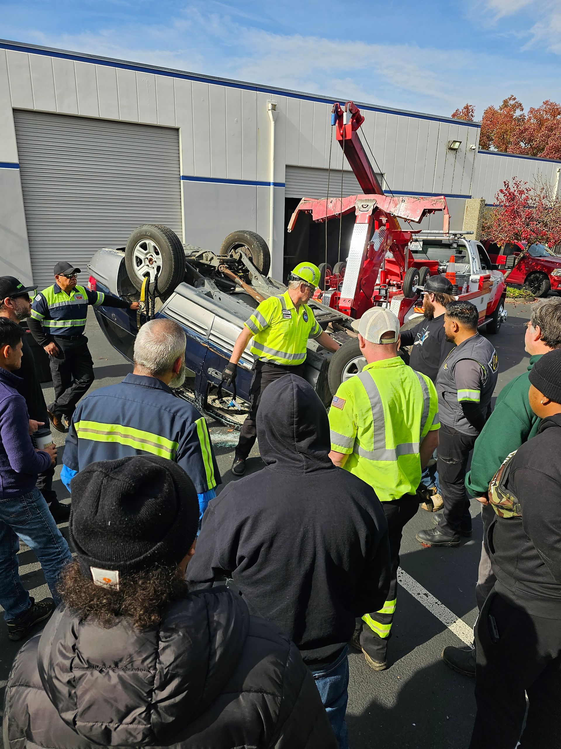 Emergency responders gather around an overturned car outside a warehouse, with a fire truck and ladder nearby.