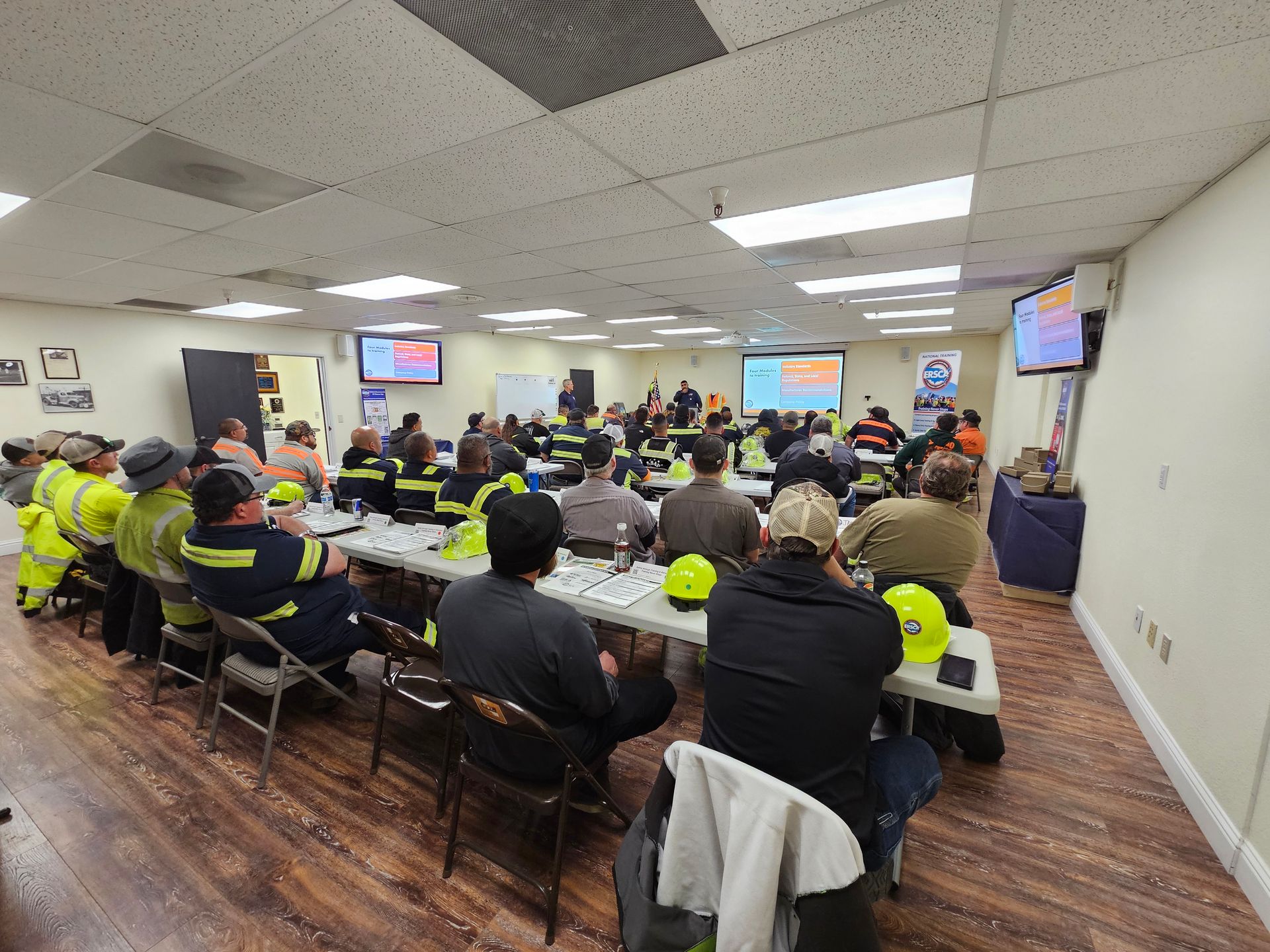 Emergency training briefing in a large room, with seated attendees watching presentation screens.
