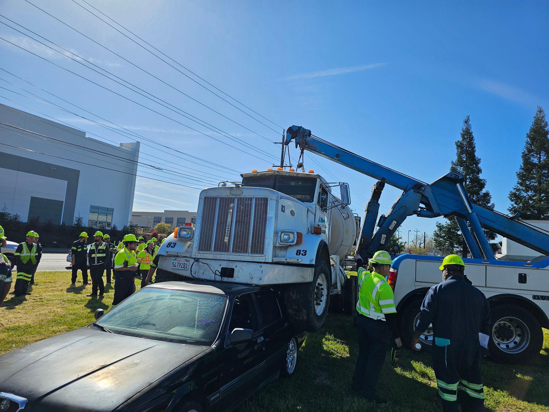 Tow truck lifting a white truck off a black car while workers in safety vests stand nearby outdoors
