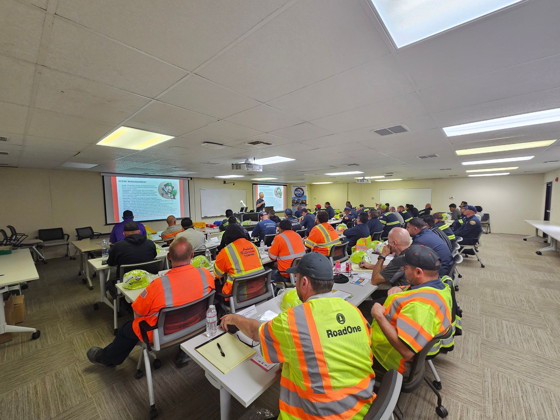 People in reflective vests attend a meeting in a large training room with tables and a presentation screen