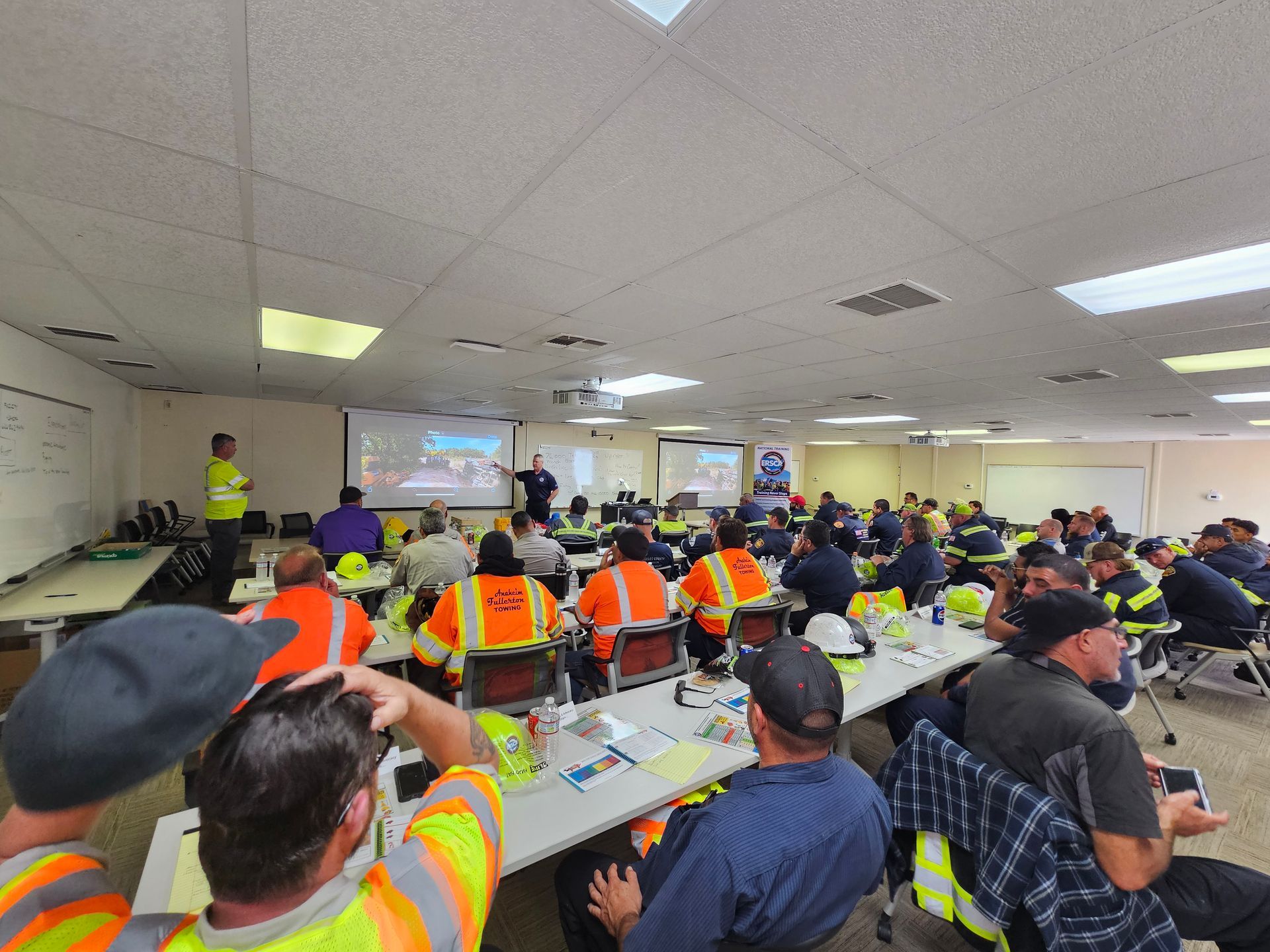 Workers in safety vests seated in a meeting room, watching a presentation at the front.