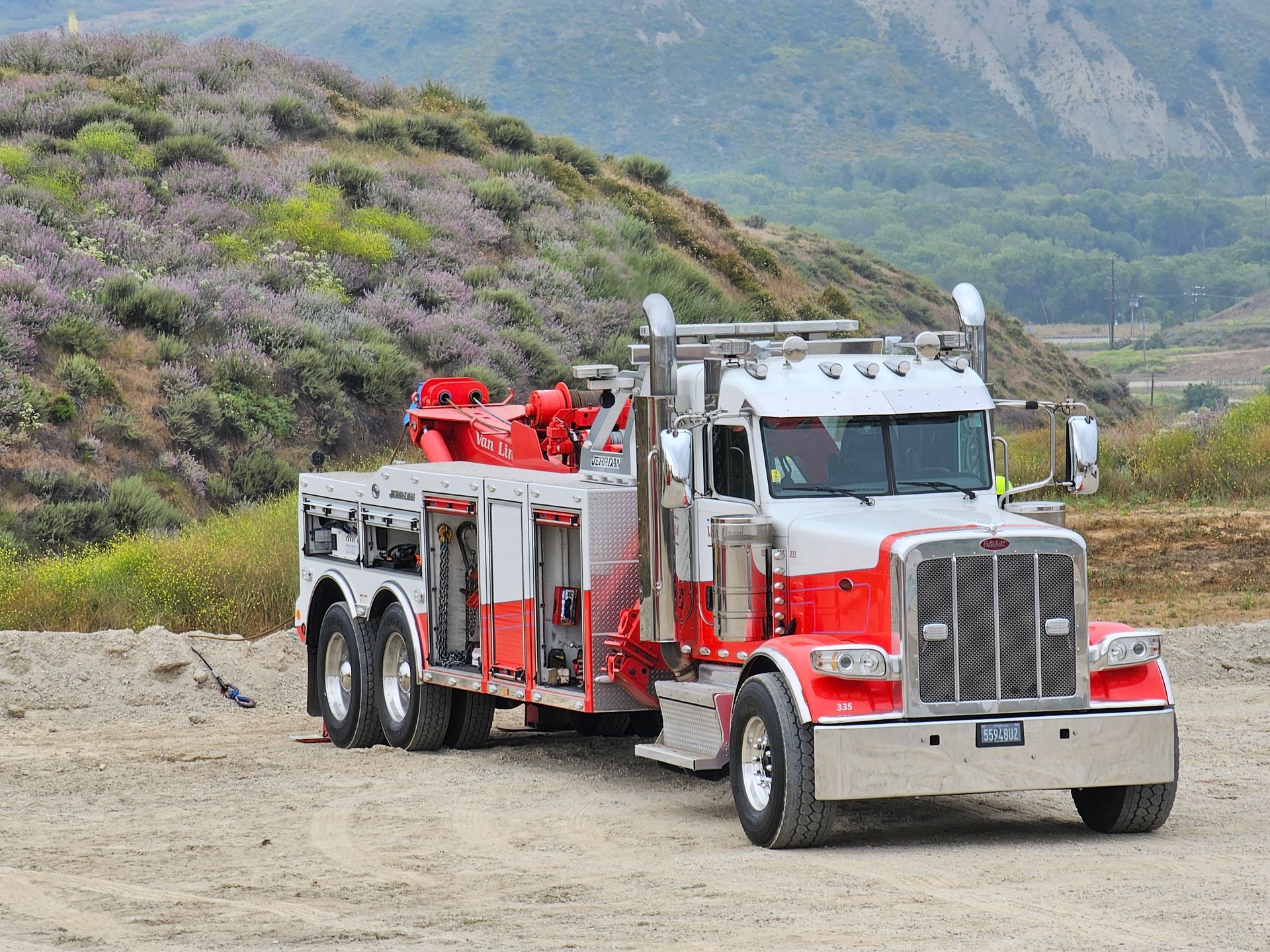 Red semi-truck with equipment driving on a dirt road beside a hillside.