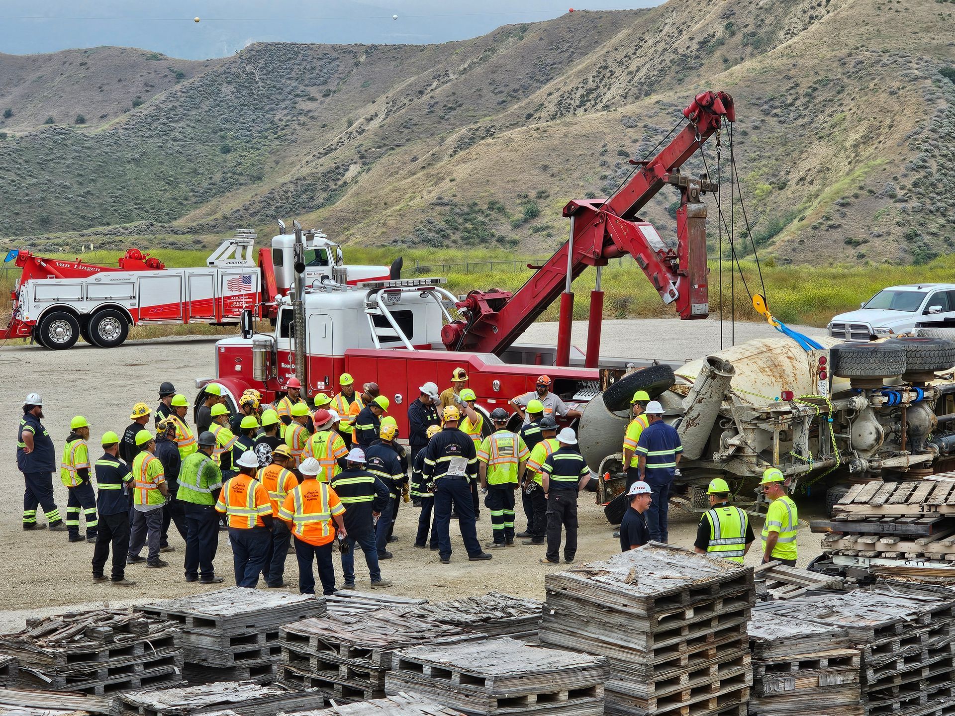 Fire crews and workers gather beside a red tow truck and crushed vehicles in a roadside mountain recovery scene
